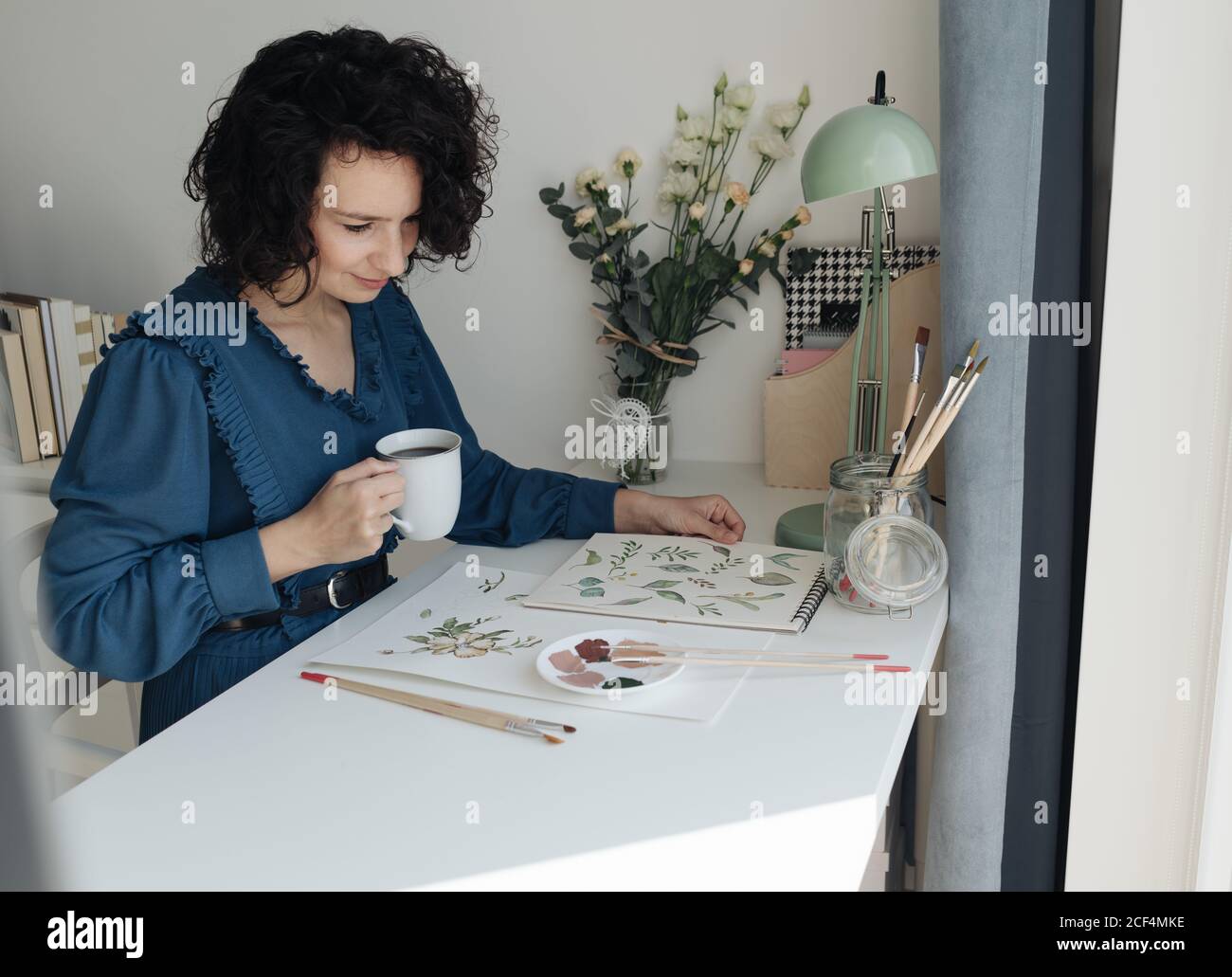 Brunette femme artiste assis à table avec une tasse de café et regardant des dessins en milieu de travail Banque D'Images