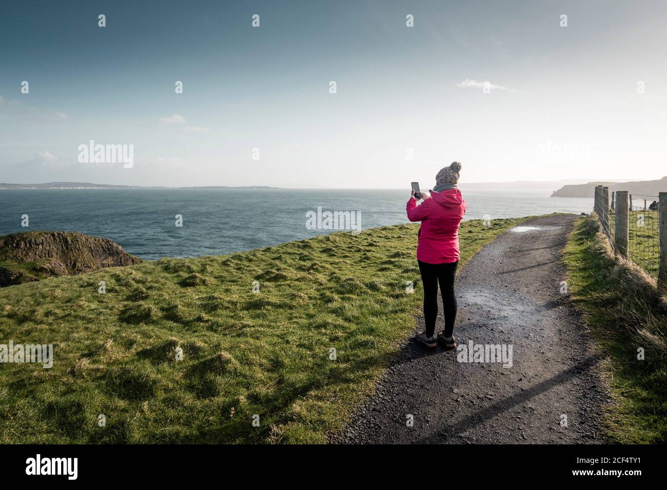 Vue arrière de la femme voyageur dans une chaude activewear debout route rurale longeant le rivage avec de l'herbe verte et de prendre photo de l'océan avec de l'eau calme le jour ensoleillé du printemps En Irlande du Nord Banque D'Images