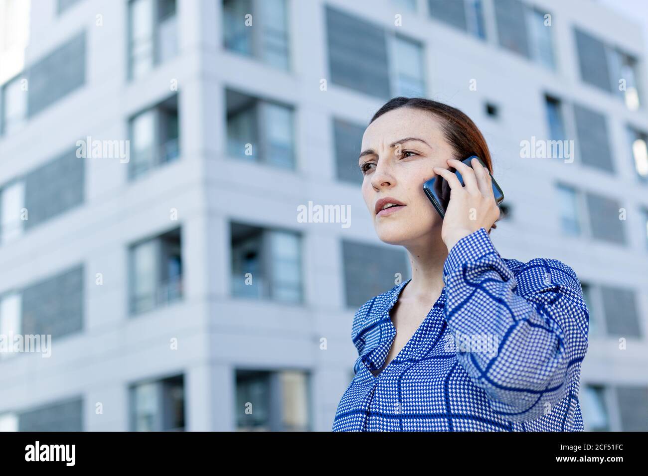 Belle femme d'affaires en chemise à carreaux bleus frognant et regardant loin tout en se tenant sur un fond flou de bâtiment moderne sur la rue de la ville Banque D'Images