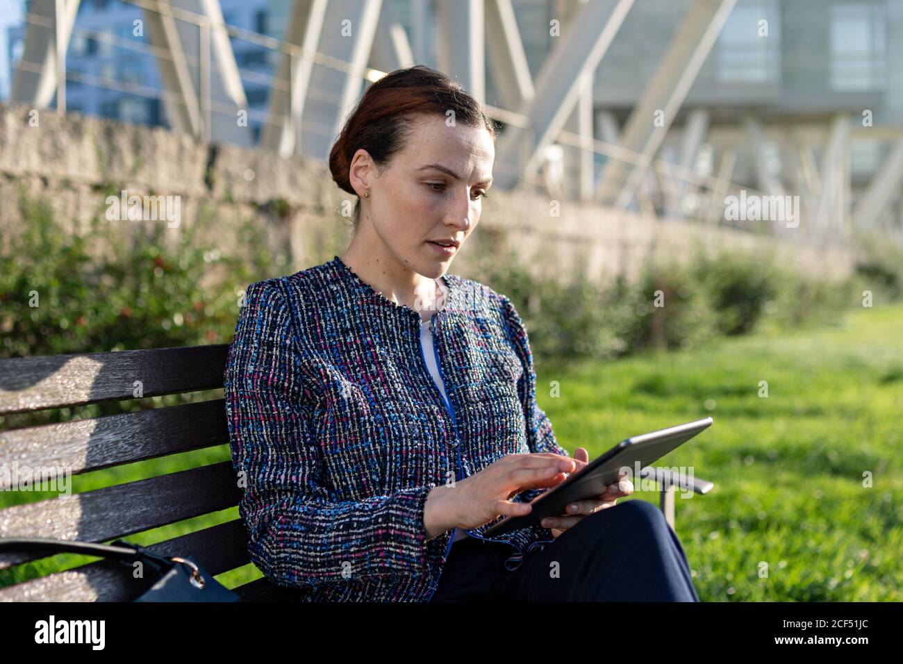 Femme entrepreneure sérieuse dans une veste élégante frowning et utilisant une tablette assis sur un banc, par beau temps, dans le parc de la ville Banque D'Images