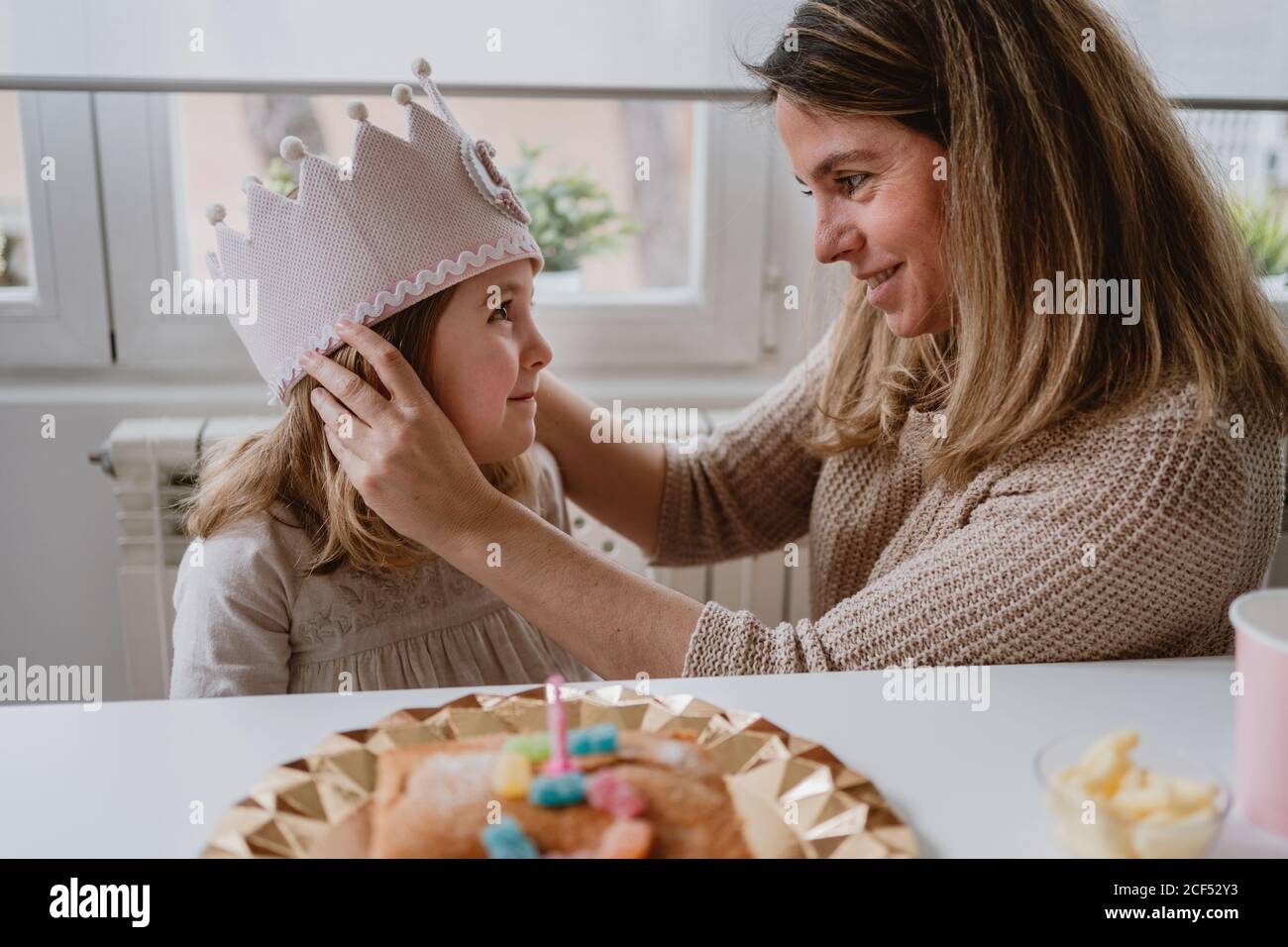 Vue latérale d'une mère gaie en plaçant une couronne faite à la main fille tout en célébrant ensemble l'anniversaire à la maison Banque D'Images Vue latérale d'une mère gaie en plaçant une couronne faite à la main fille tout en célébrant ensemble l'anniversaire à la maison Banque D'Images