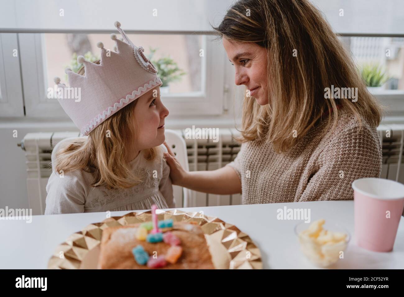 Vue latérale d'une mère gaie en plaçant une couronne faite à la main fille tout en célébrant ensemble l'anniversaire à la maison Banque D'Images Vue latérale d'une mère gaie en plaçant une couronne faite à la main fille tout en célébrant ensemble l'anniversaire à la maison Banque D'Images