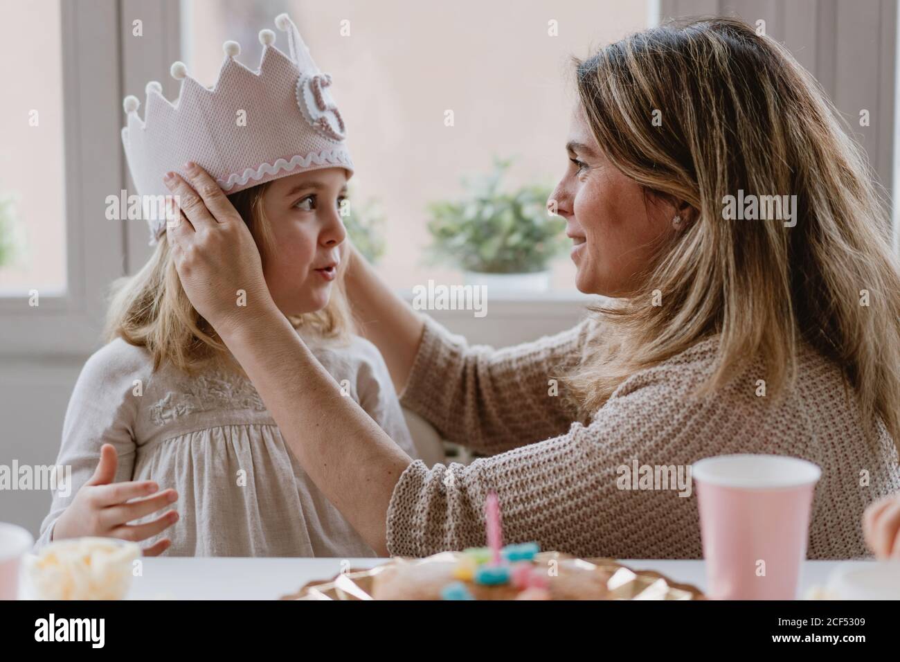 Vue latérale d'une mère gaie en plaçant une couronne faite à la main fille tout en célébrant ensemble l'anniversaire à la maison Banque D'Images Vue latérale d'une mère gaie en plaçant une couronne faite à la main fille tout en célébrant ensemble l'anniversaire à la maison Banque D'Images