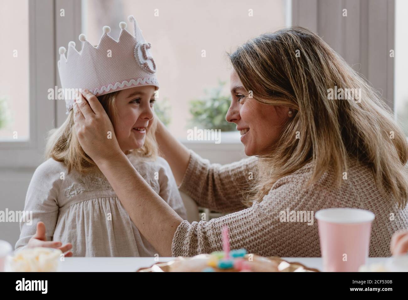 Vue latérale d'une mère gaie en plaçant une couronne faite à la main fille tout en célébrant ensemble l'anniversaire à la maison Banque D'Images Vue latérale d'une mère gaie en plaçant une couronne faite à la main fille tout en célébrant ensemble l'anniversaire à la maison Banque D'Images