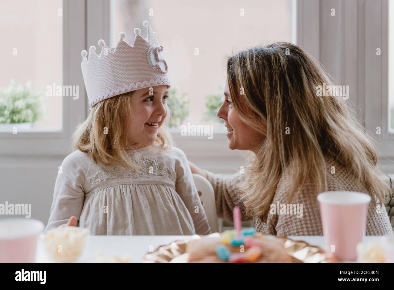 Vue latérale d'une mère gaie en plaçant une couronne faite à la main fille tout en célébrant ensemble l'anniversaire à la maison Banque D'Images Vue latérale d'une mère gaie en plaçant une couronne faite à la main fille tout en célébrant ensemble l'anniversaire à la maison Banque D'Images