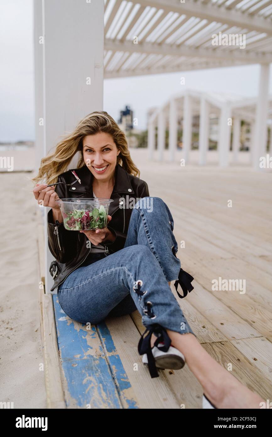 Élégante femme blonde aux cheveux longs en blouson noir appréciant une salade verte saine tout en étant assise sur une terrasse en bois sur la côte en souriant à l'appareil photo Banque D'Images