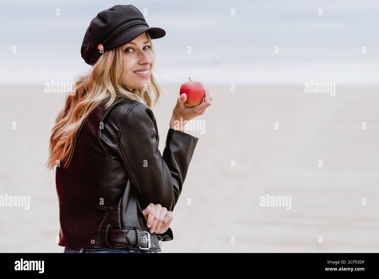 Vue arrière de la femme blonde tendance en casquette noire et veste en cuir  avec gaiement sur l'épaule avec pomme mûre rouge à la main sur la plage de  sable Photo Stock -