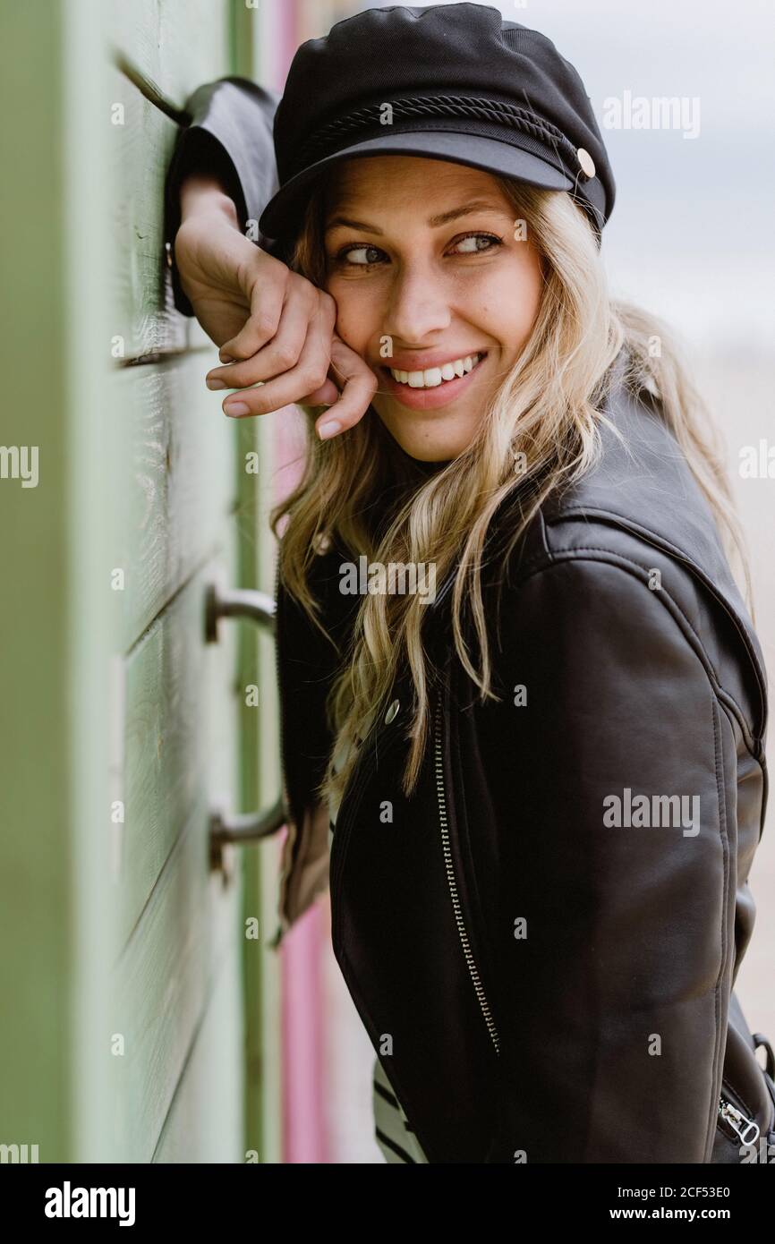 Femme blonde aux cheveux longs tendance en casquette noire et veste en cuir  souriant aux couleurs vives et penchée sur le mur des cabines de plage en  bois Photo Stock - Alamy
