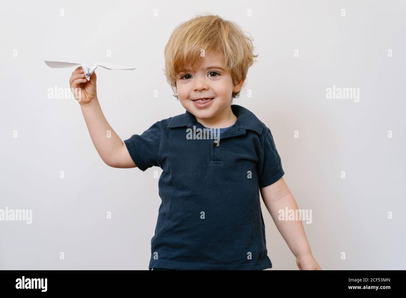 Joli garçon blond dans un ensemble décontracté jouant avec un rabot en papier sourire éclatant à l'appareil photo sur fond de mur blanc Banque D'Images