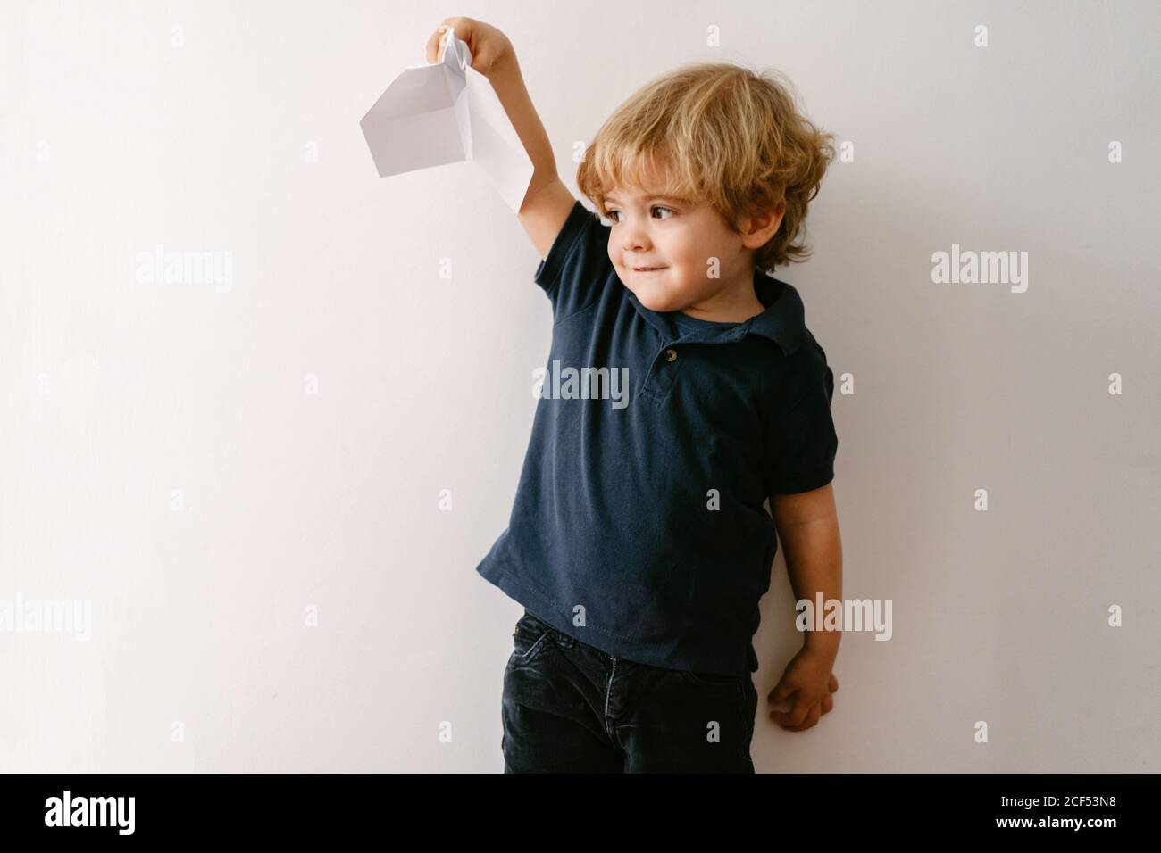 Joli garçon blond dans un ensemble décontracté jouant avec un rabot en papier sourire éclatant à l'appareil photo sur fond de mur blanc Banque D'Images