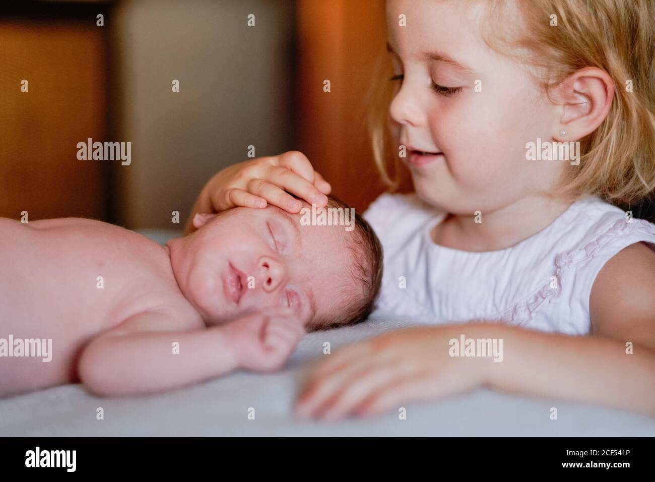 Joyeuse petite récolte préscolaire femelle touchant avec intérêt calme bébé avec les yeux fermés reposant dans le lit contre l'intérieur flou de chambre lumineuse Banque D'Images