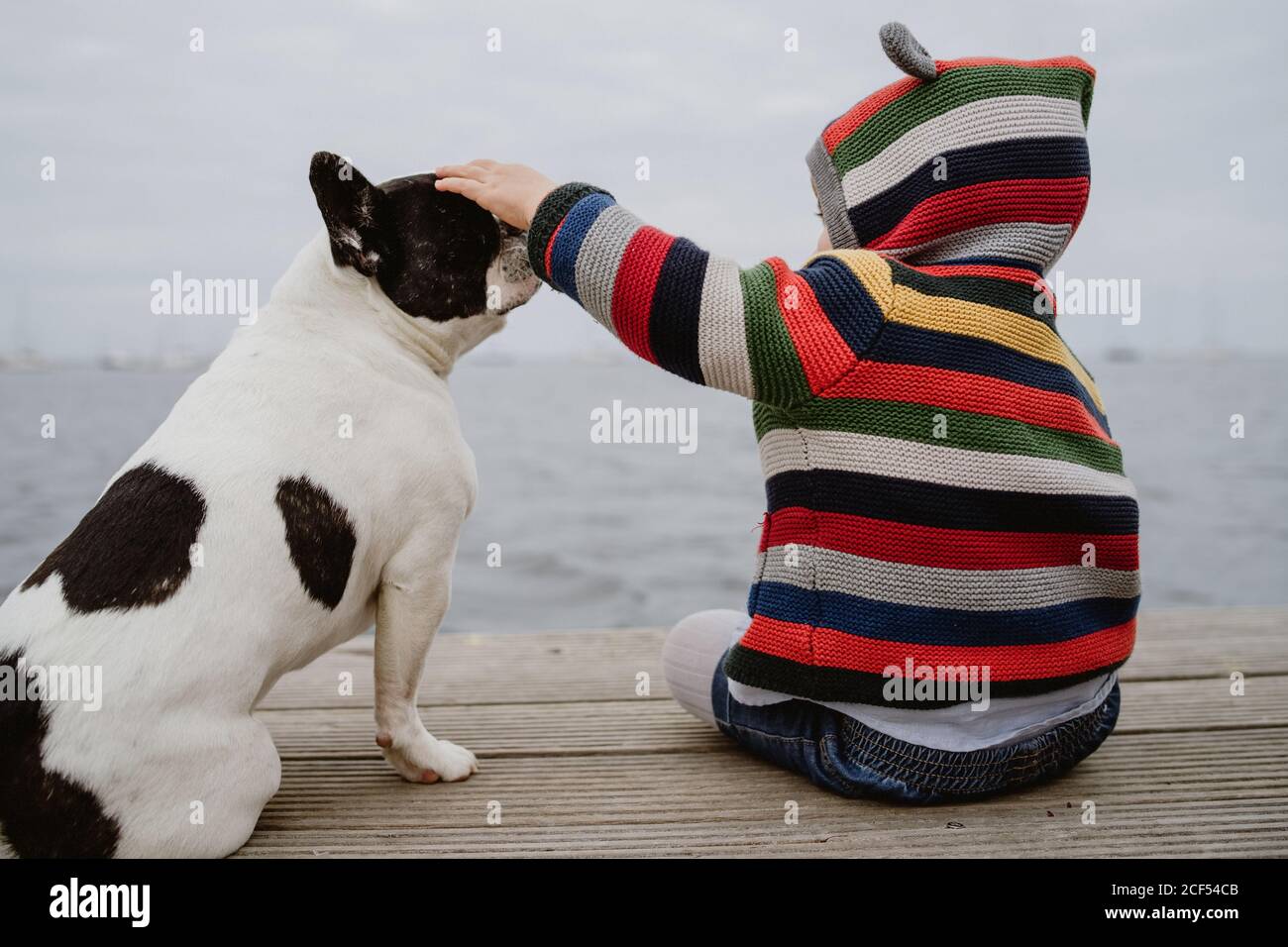 Vue arrière d'un enfant méconnaissable dans une veste rayée à pois Bulldog français assis sur une jetée près de la mer Banque D'Images