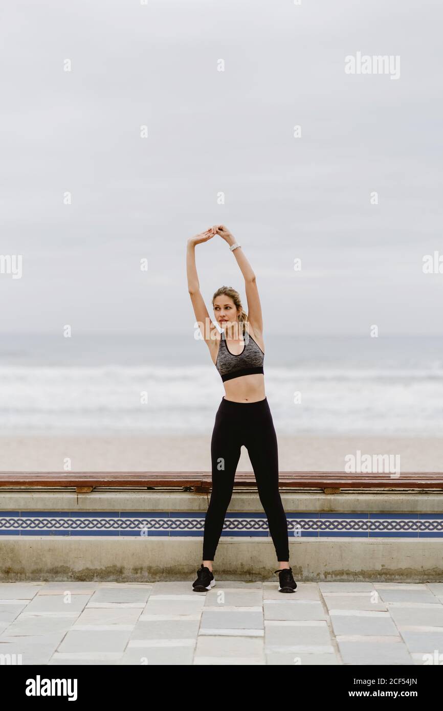 Athlète femme en tenue active debout avec les mains levées dans la serrure et s'étirant près du bord de mer par temps nuageux Banque D'Images