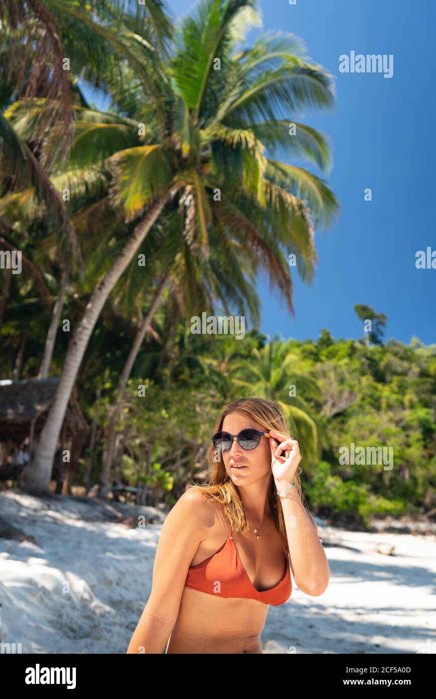 Jeune femme en maillot de bain, bain de soleil sur un bord de mer sablonneux sur fond de palmiers tropicaux et de ciel bleu Banque D'Images