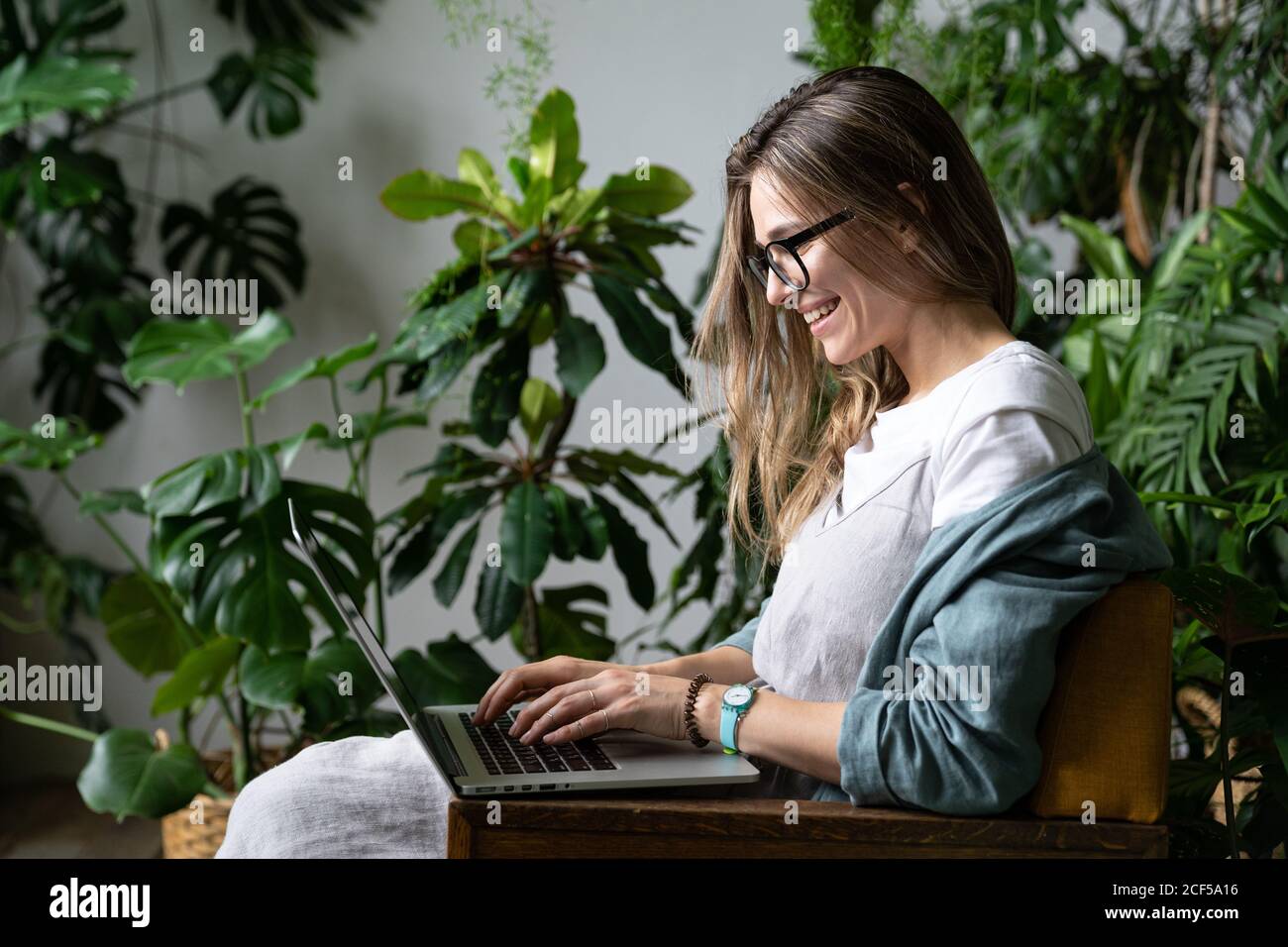 Gros plan portrait d'une femme souriante gardenier en lunettes porter une robe de lin, s'asseoir sur une chaise dans une maison verte, utiliser un ordinateur portable et parler sur Internet Banque D'Images