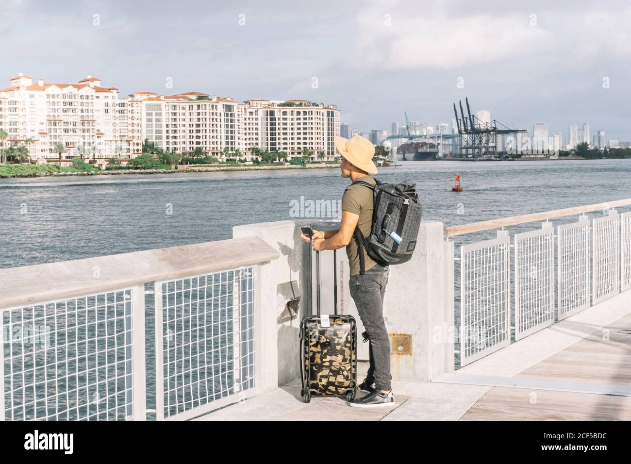 Jeune homme avec une valise et un sac à dos, arrivant sur les rives de Miami, regardant la rivière et les bâtiments un matin ensoleillé Banque D'Images