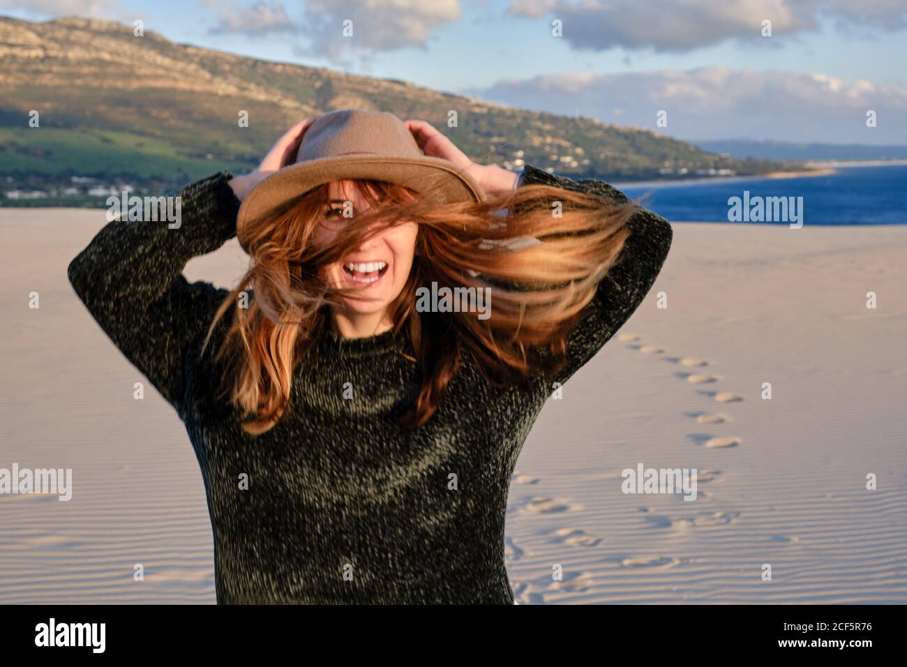 Joyeuse femme de voyage en chapeau debout dans un désert de sable isolé au coucher du soleil regardant la caméra à Tarifa, Espagne Banque D'Images