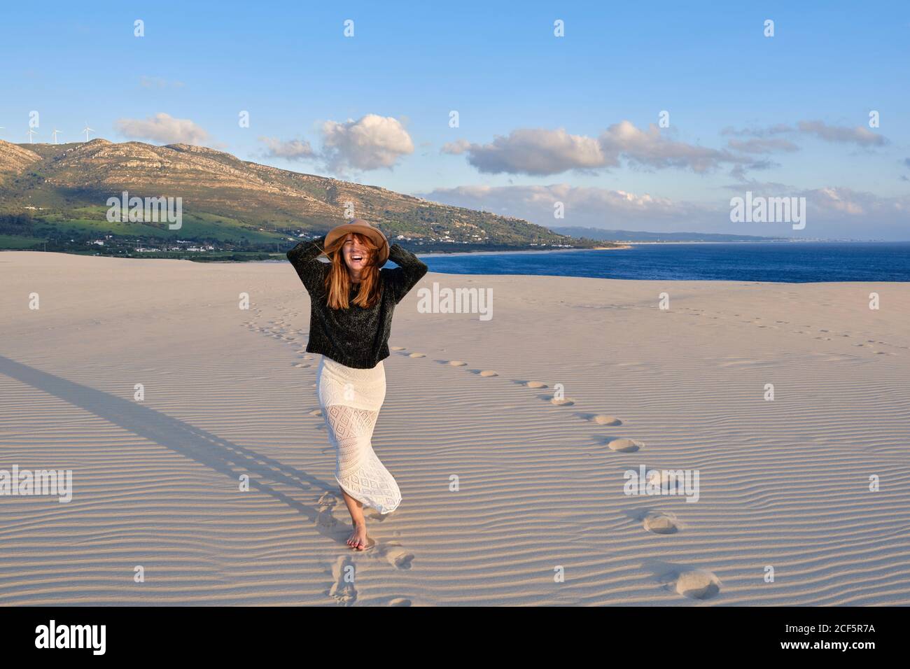 Joyeuse femme de voyage en chapeau debout dans un désert de sable isolé au coucher du soleil regardant la caméra à Tarifa, Espagne Banque D'Images