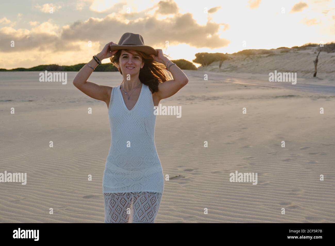Joyeuse femme de voyage en chapeau debout dans un désert de sable isolé au coucher du soleil regardant la caméra à Tarifa, Espagne Banque D'Images