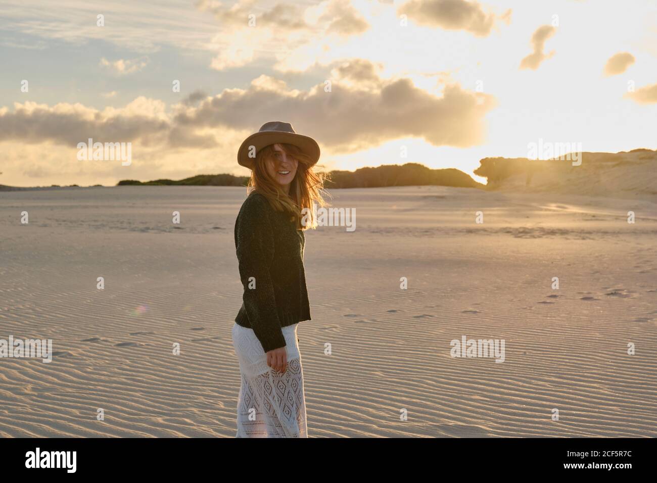 Joyeuse femme de voyage en chapeau debout dans un désert de sable isolé au coucher du soleil regardant la caméra à Tarifa, Espagne Banque D'Images