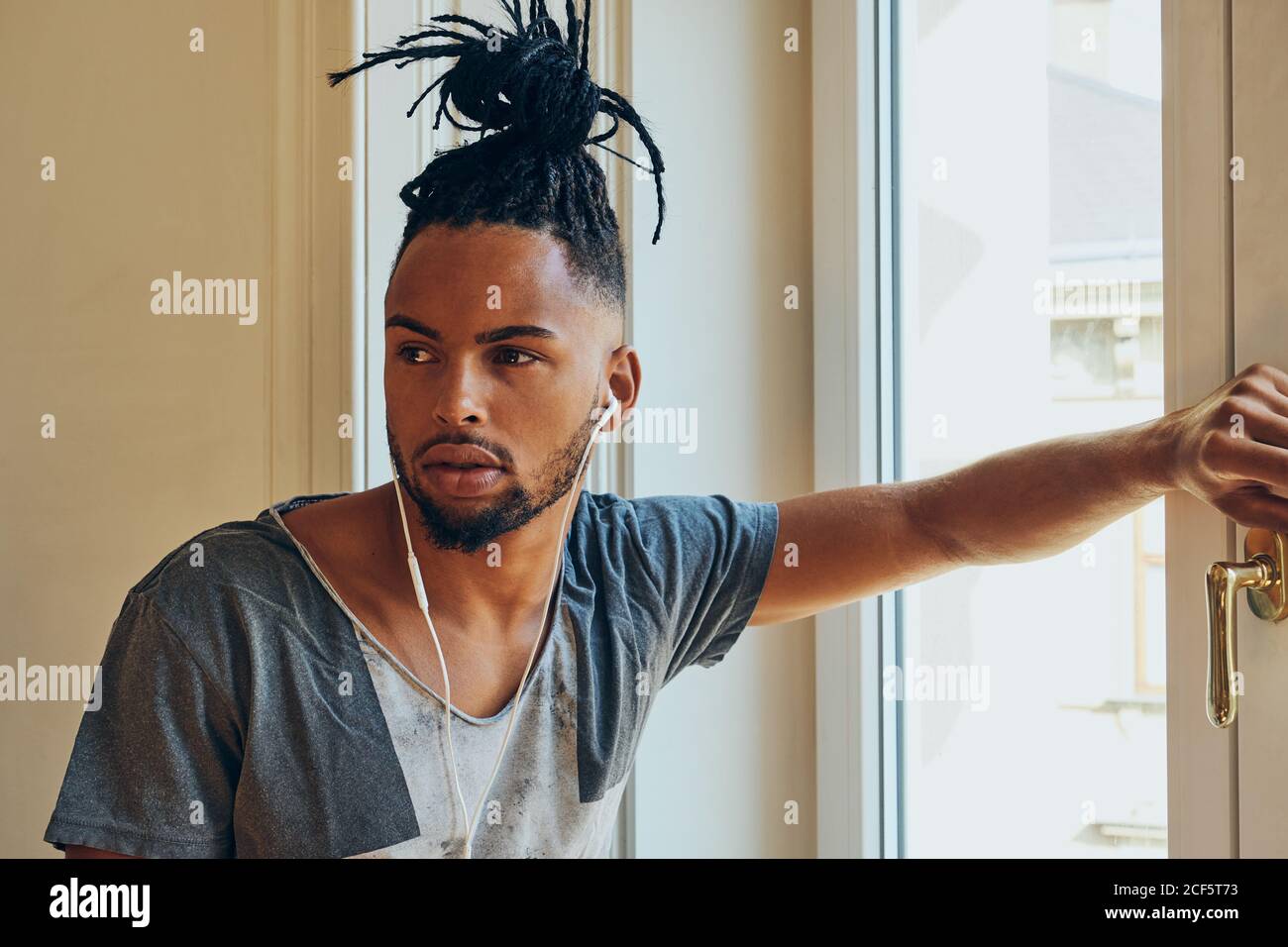 Vue latérale d'un homme ethnique sérieux avec une coiffure créative à l'écoute pour écouter de la musique et vous pencher sur le cadre de la fenêtre Banque D'Images