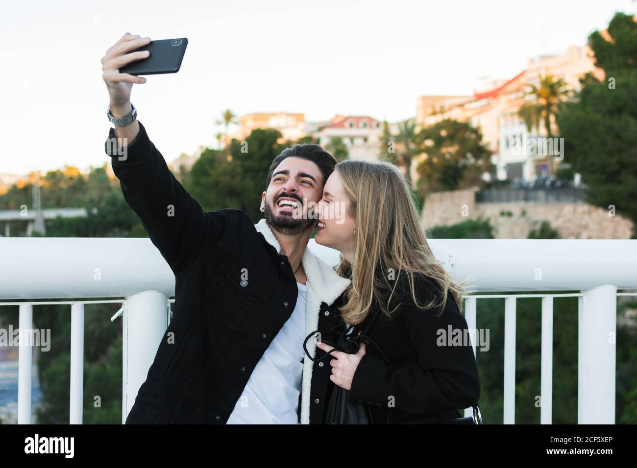Joyeux jeune couple multiracial dans une tenue décontractée prenant le selfie sur téléphone portable en se tenant ensemble sur un pont avec des arbres verts et les bâtiments de la ville en arrière-plan Banque D'Images