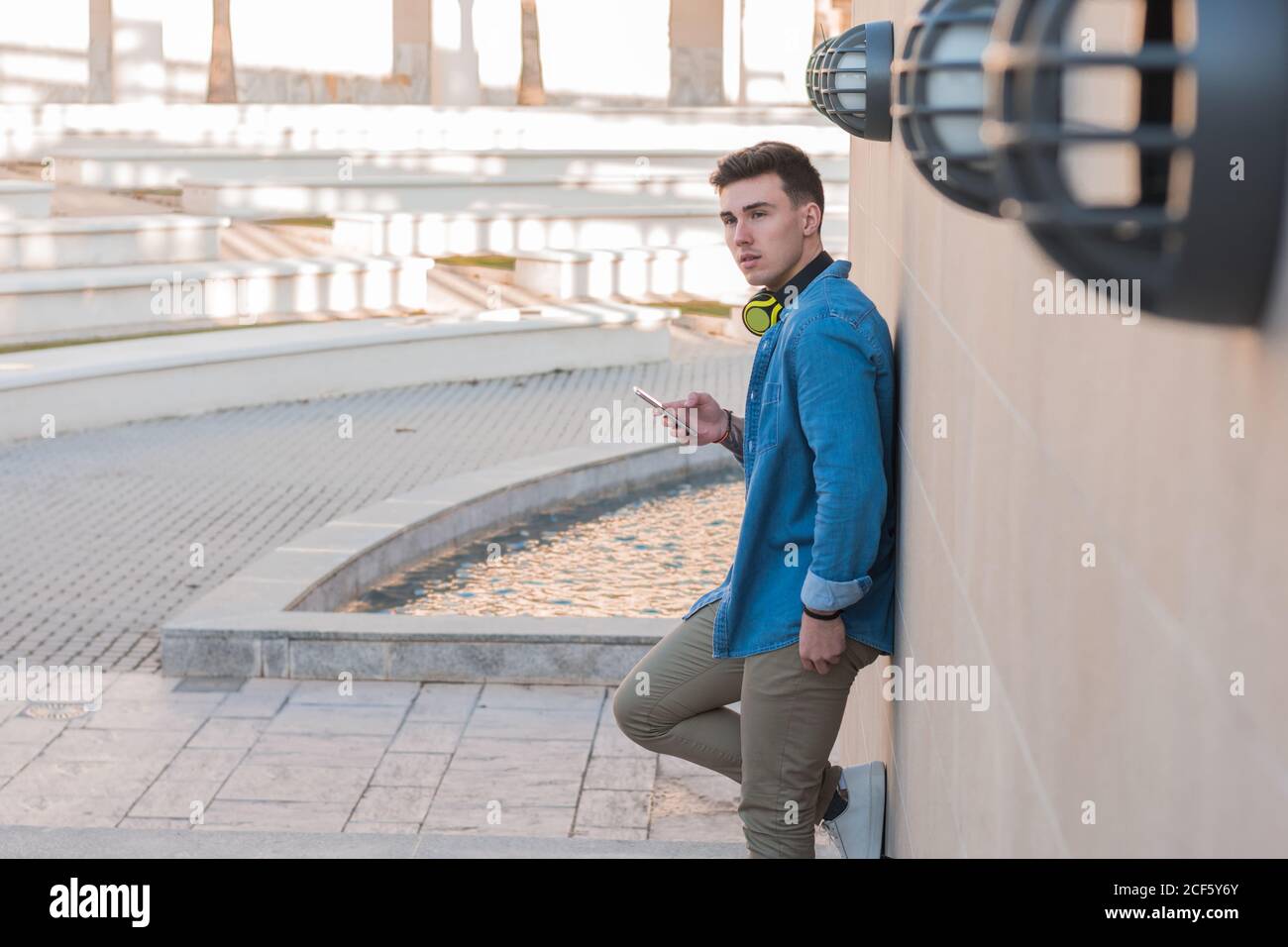 Vue latérale d'un homme élégant et élégant dans un casque de surf lumineux téléphone mobile tout en s'appuyant sur un mur en marbre par beau temps Banque D'Images