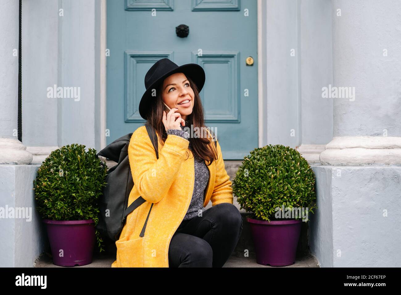 Femme gaie dans un manteau et un chapeau jaune élégant souriant et  regardant loin tout en étant assis près des plantes en pot et porte et  répondant à un appel téléphonique dans