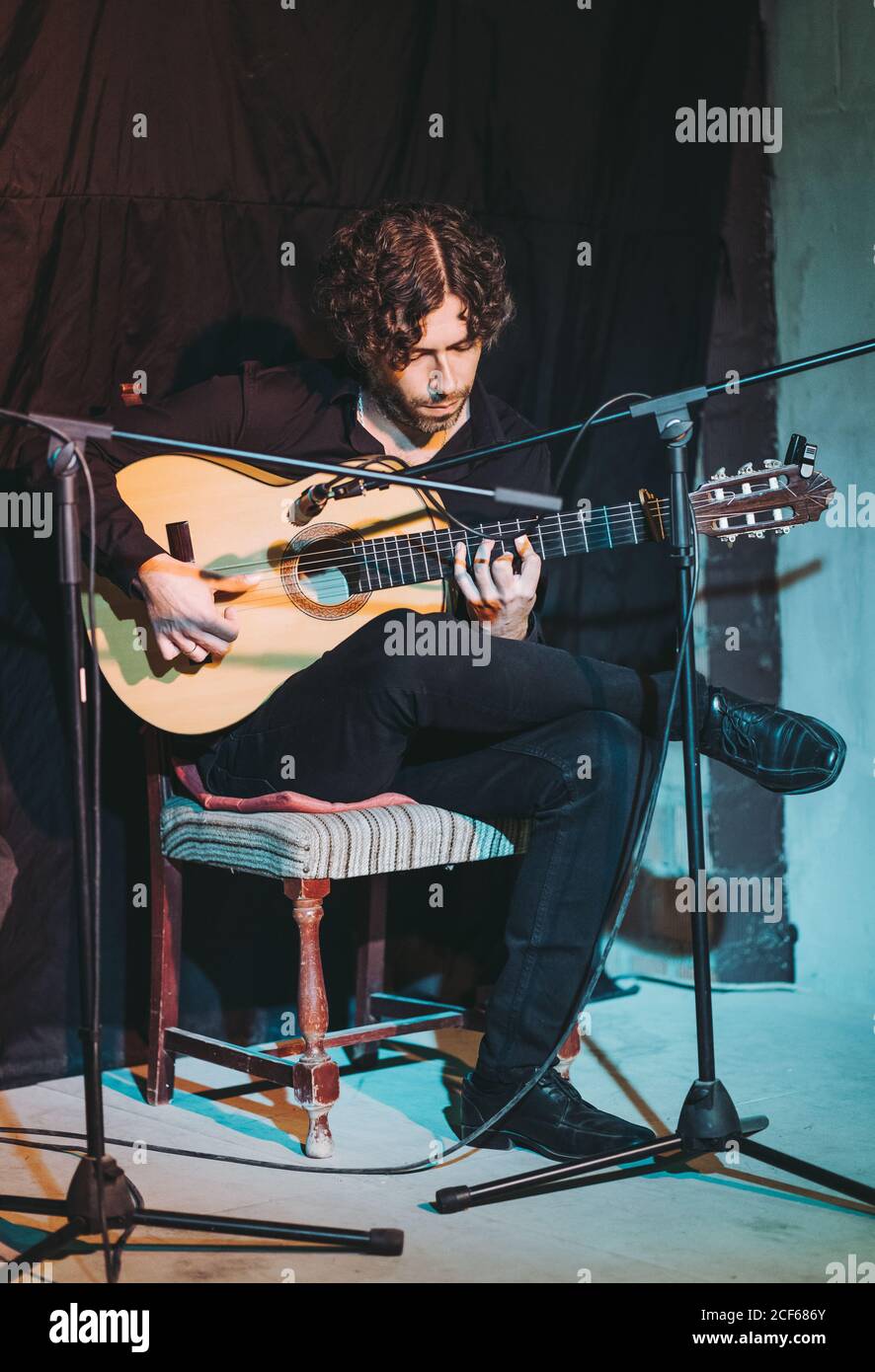 Musicien hispanique en noir assis avec les jambes croisées chaise en bois près du microphone tout en jouant de la guitare sur la plate-forme de théâtre Banque D'Images