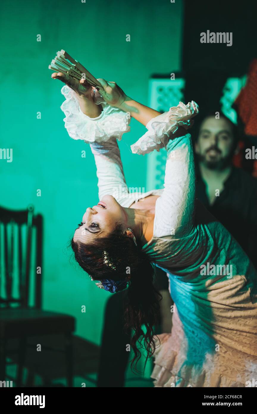 Danseuse hispanique adulte en robe rouffée prenant un fan de main tout en dansant avec les bras levés devant le musicien barbu pendant la performance Banque D'Images