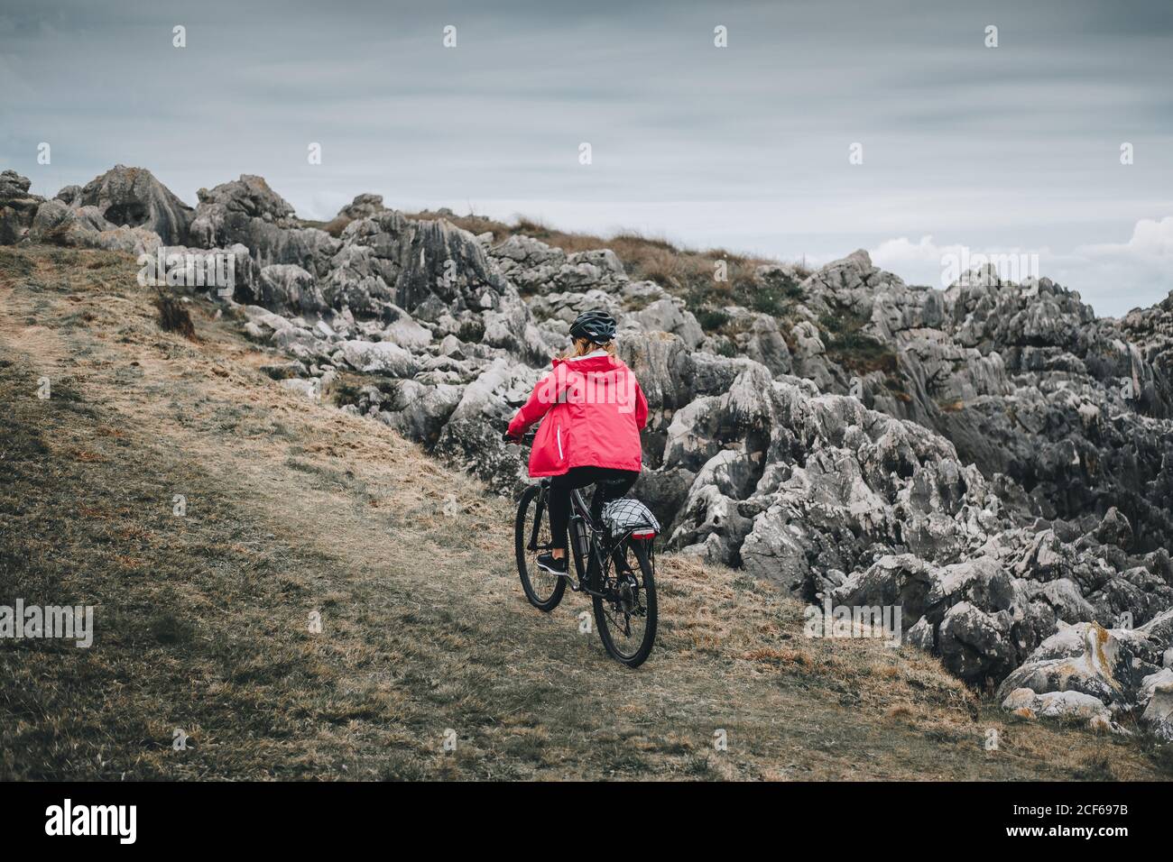Vue arrière de la femme anonyme en casque noir et veste rouge vélo sur route rocheuse déserte le jour nuageux Banque D'Images