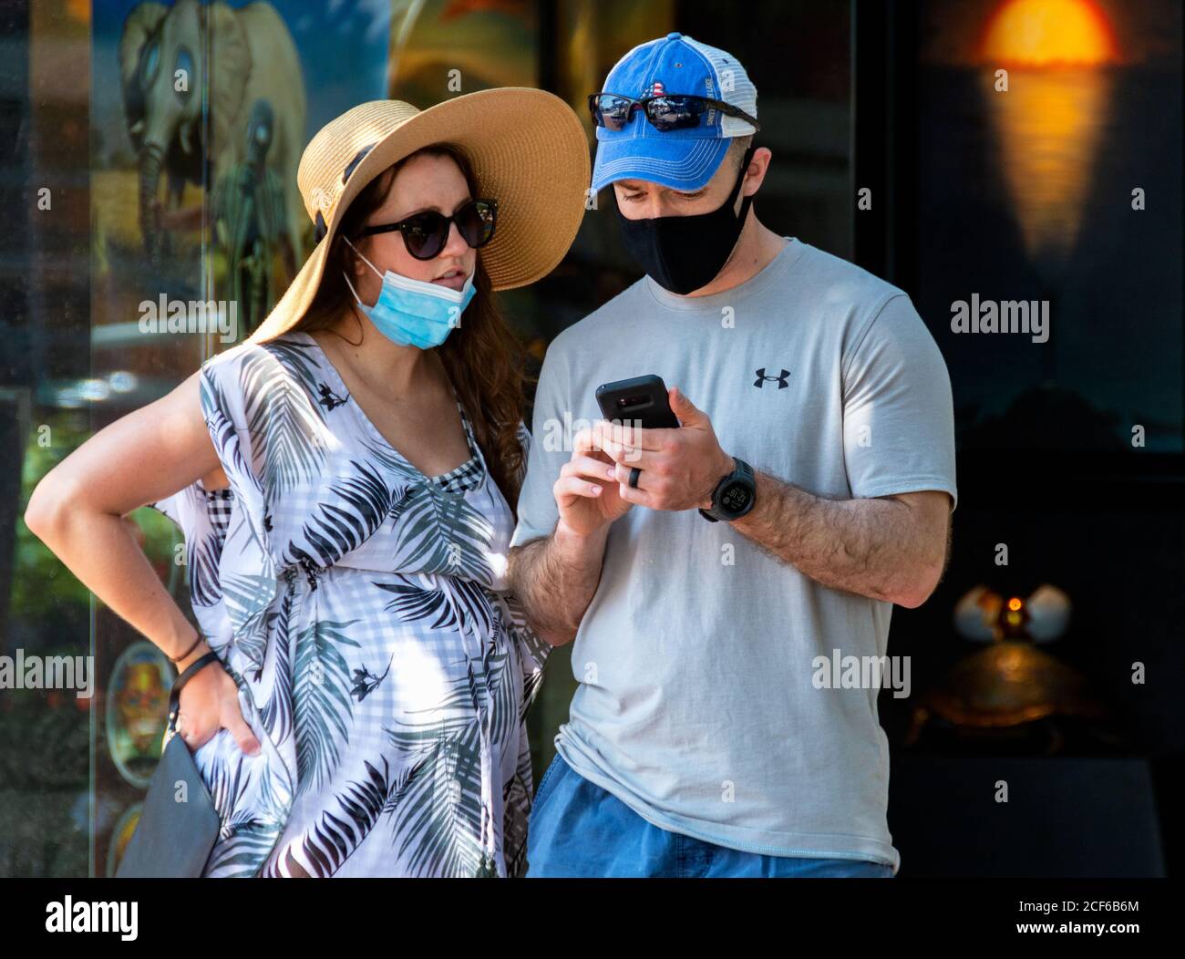 Femme enceinte, portant un masque facial de protection sur son menton, regarde comme mari, portant un masque facial correctement, utilise un smartphone en face de la boutique. Banque D'Images