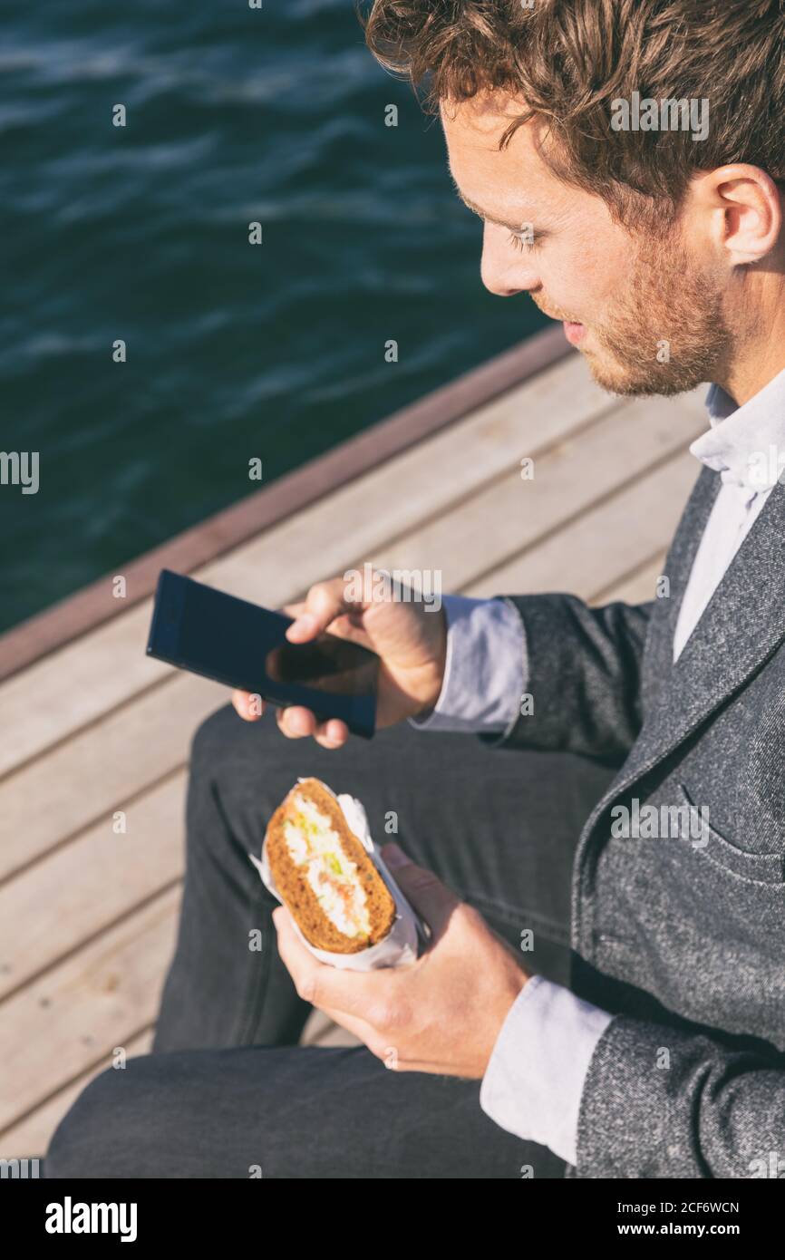 Homme professionnel en pause déjeuner de travail en utilisant le téléphone portable manger un sandwich à l'extérieur près du canal d'eau. Jeune adulte du Caucase tenant un smartphone mobile Banque D'Images