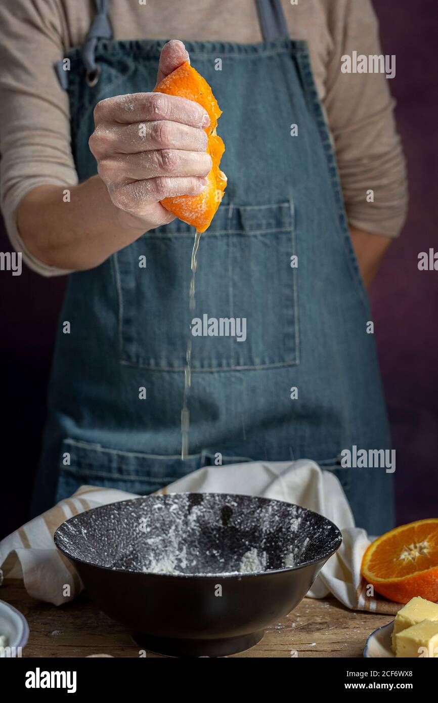 Rognez la main d'une femme méconnaissable dans un tablier en pressant l'orange fraîchement coupée sur un bol tout en préparant la pâte à la table Banque D'Images