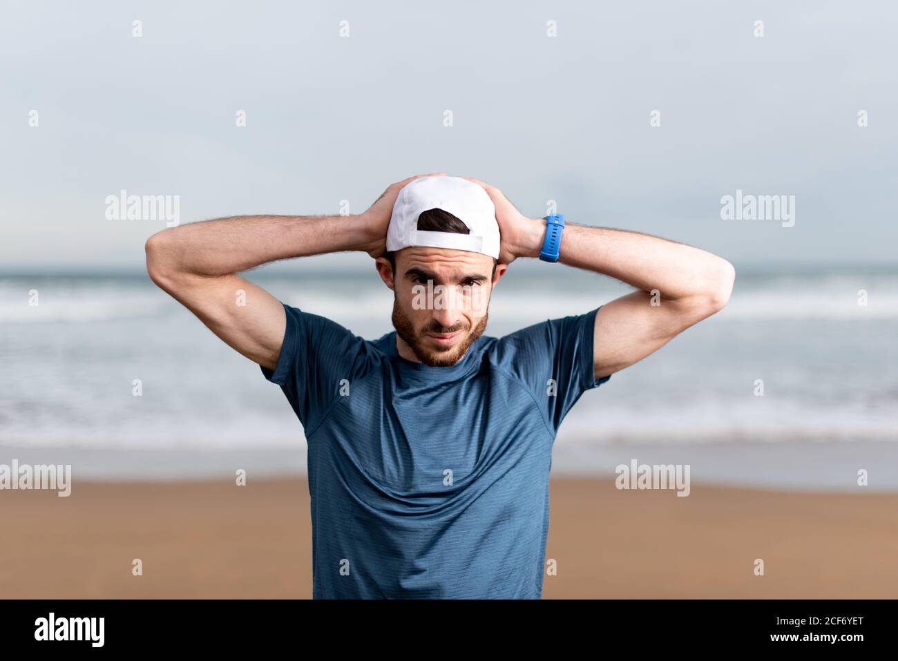 Sportsman en t-shirt bleu avec les mains derrière la tête casquette blanche  regardant l'appareil photo avec sable blanc sur la mer arrière-plan flou  Photo Stock - Alamy