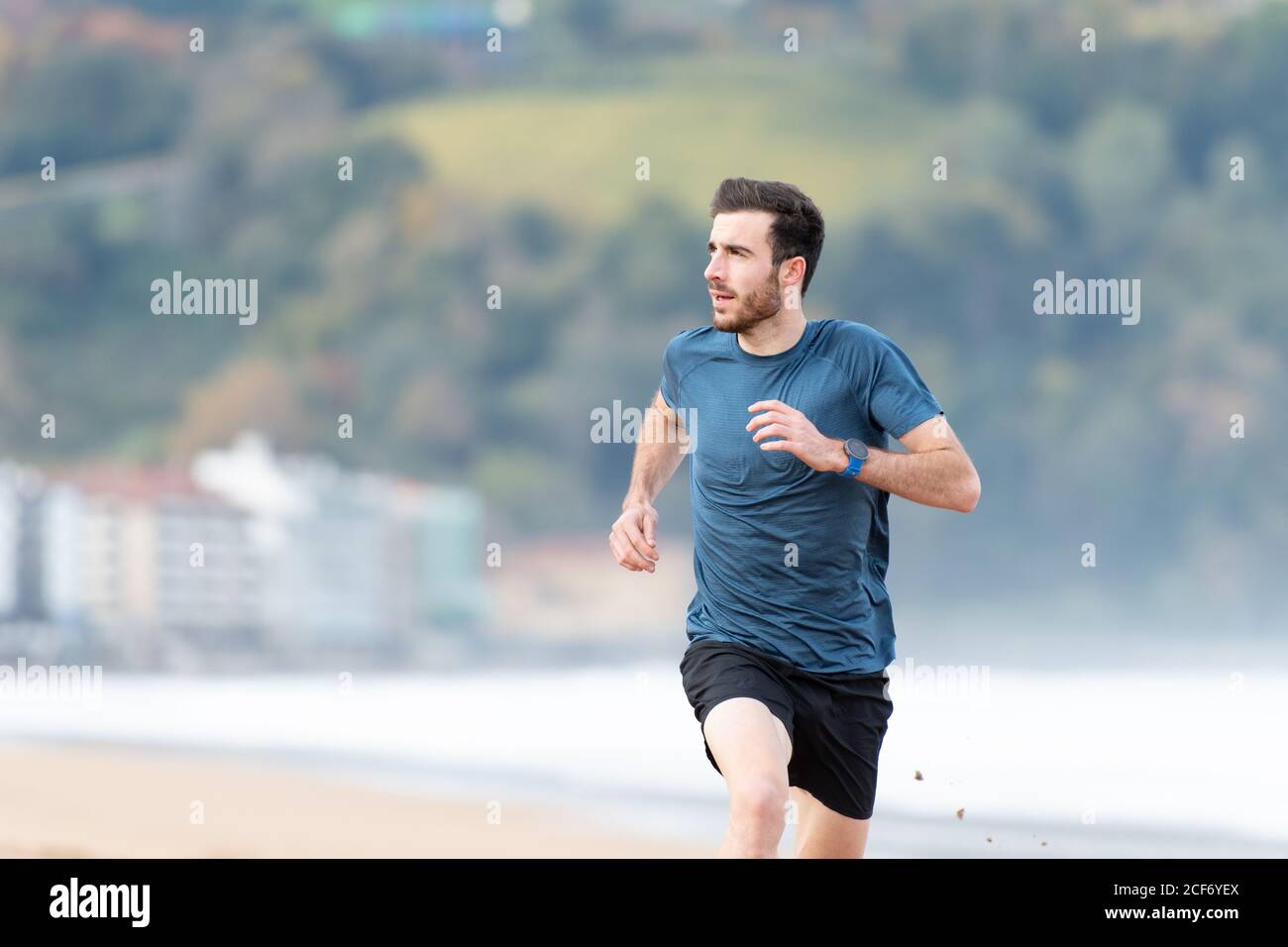 Athlète masculin barbu en tenue active en courant sur le sable vide plage avec montagnes vertes sur fond flou Banque D'Images