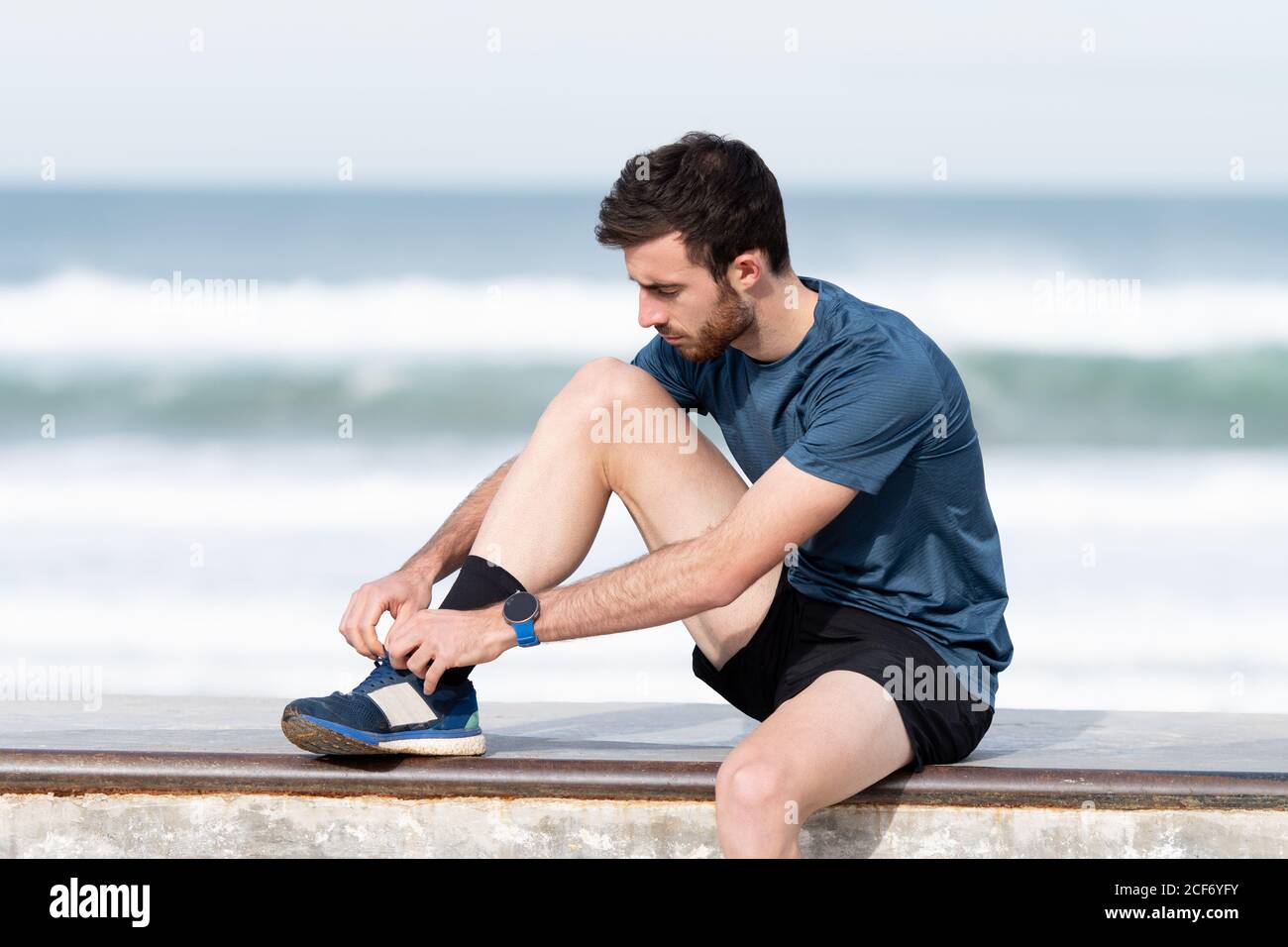 Athlète masculin à poil noir en t-shirt bleu et short assis sur la clôture et nouant les lacets avec la mer bleue et le ciel sur un arrière-plan flou Banque D'Images