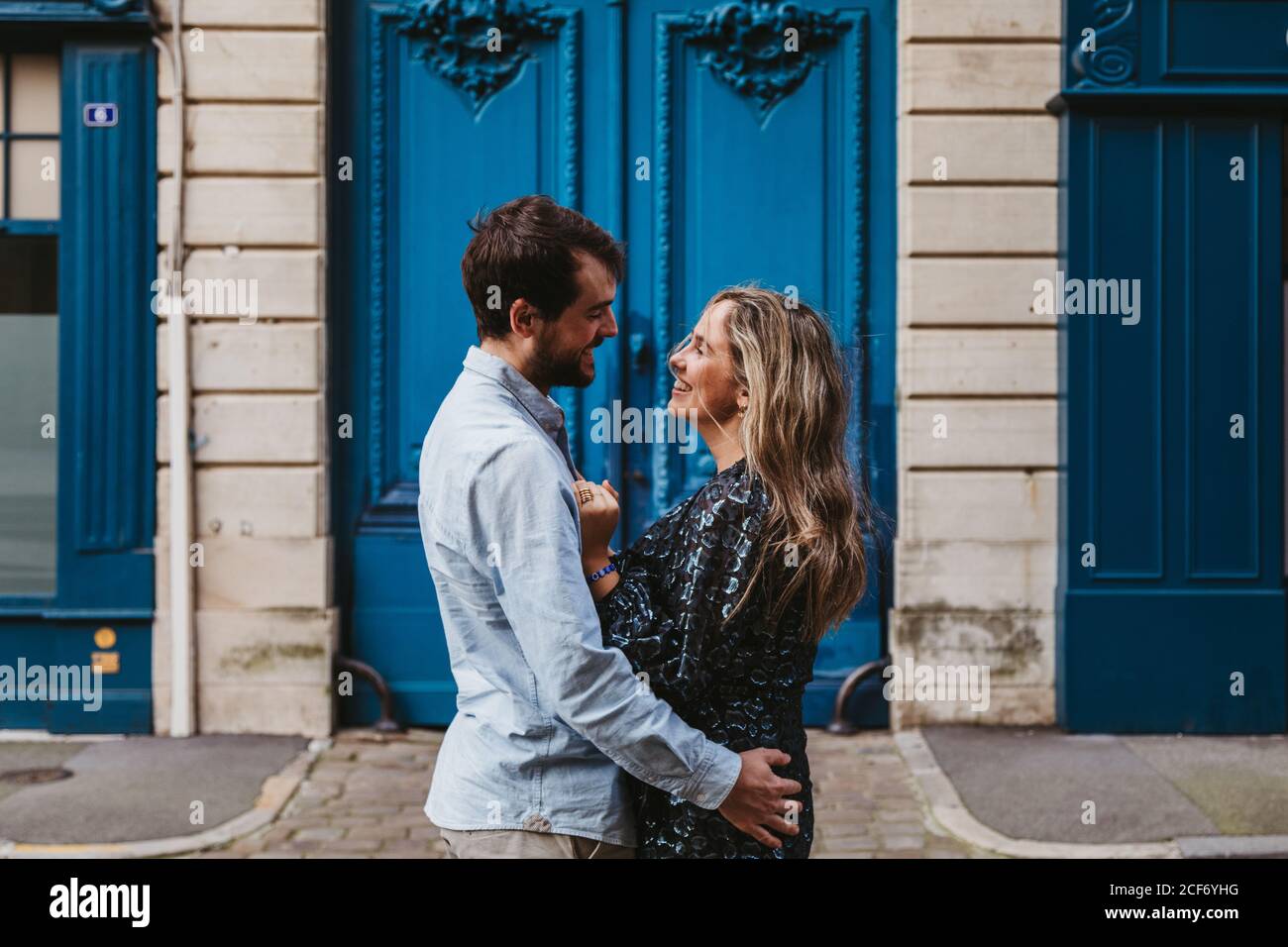 Vue latérale d'un jeune couple heureux dans des vêtements décontractés qui s'embrasent se regardant l'un contre l'autre en se tenant contre un bâtiment en pierre vieilli avec portes bleues sur la rue de la ville Banque D'Images