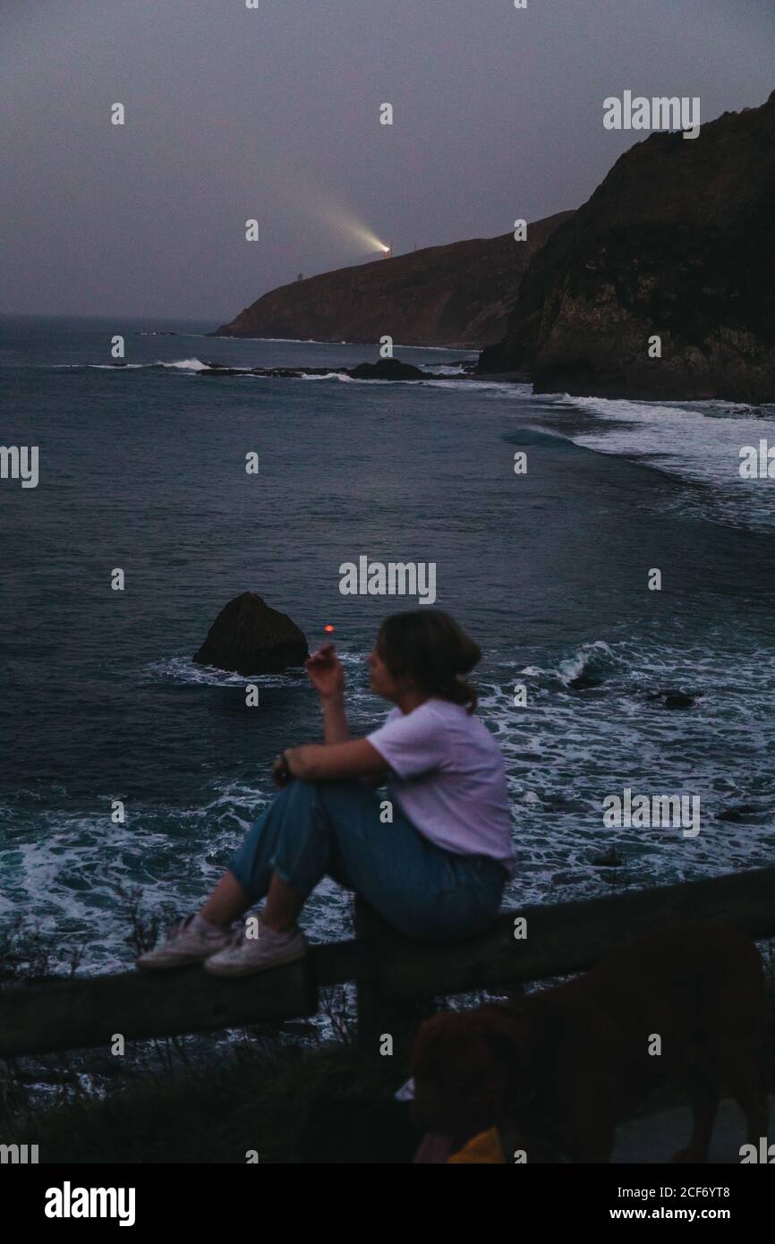 Vue d'en haut de côté de l'décontracté pensive jeune femme fumeurs cigarette assis sur la clôture sur la côte rocheuse de la mer dedans Soirée d'été dans la ville de Lekeitio en Espagne avec balise lumière en arrière-plan Banque D'Images