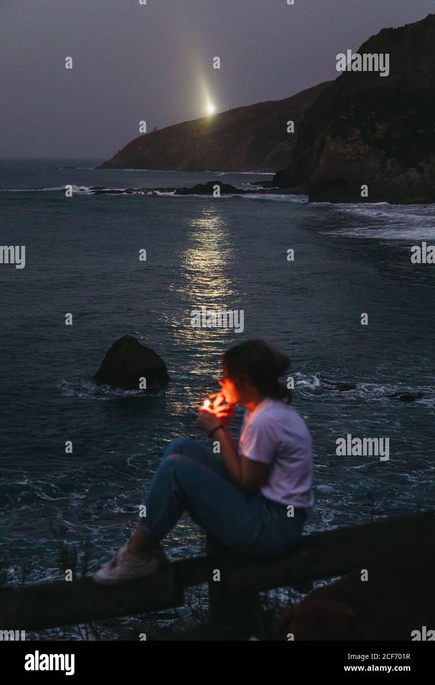 Vue d'en haut de côté de l'décontracté pensive jeune femme fumeurs cigarette assis sur la clôture sur la côte rocheuse de la mer dedans Soirée d'été dans la ville de Lekeitio en Espagne avec balise lumière en arrière-plan Banque D'Images