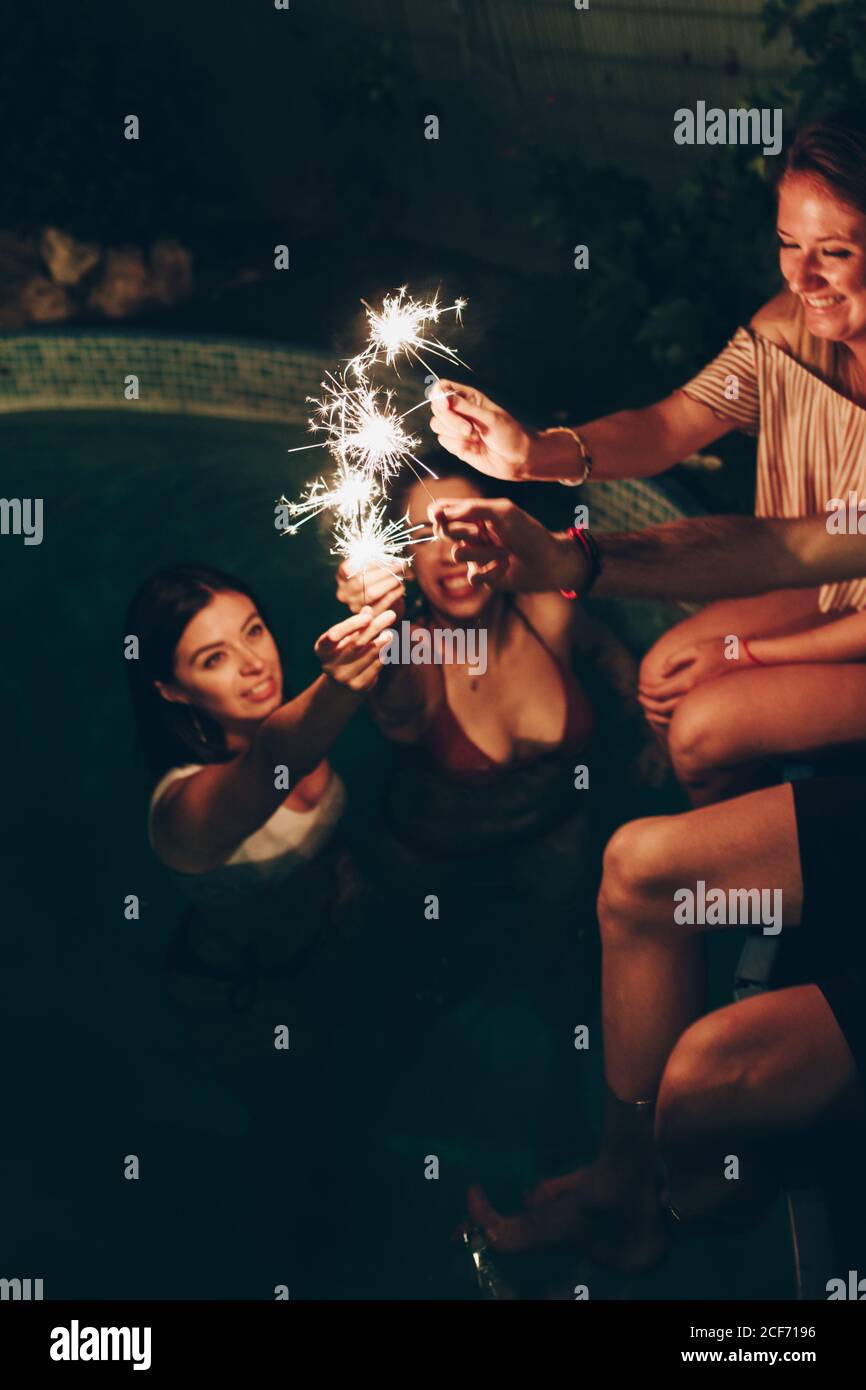 Groupe de jeunes souriant et brûlant des étincelles tout en ayant faites la fête près de la piscine la nuit Banque D'Images Groupe de jeunes souriant et brûlant des étincelles tout en ayant faites la fête près de la piscine la nuit Banque D'Images