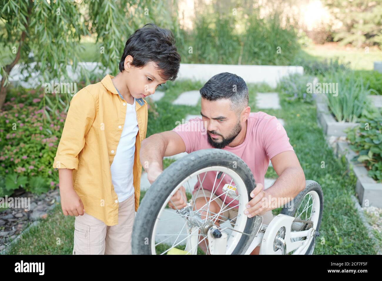 Jeune homme adulte moderne avec barbe sur le visage passant du temps à l'extérieur, aider son fils à fixer la roue de vélo Banque D'Images