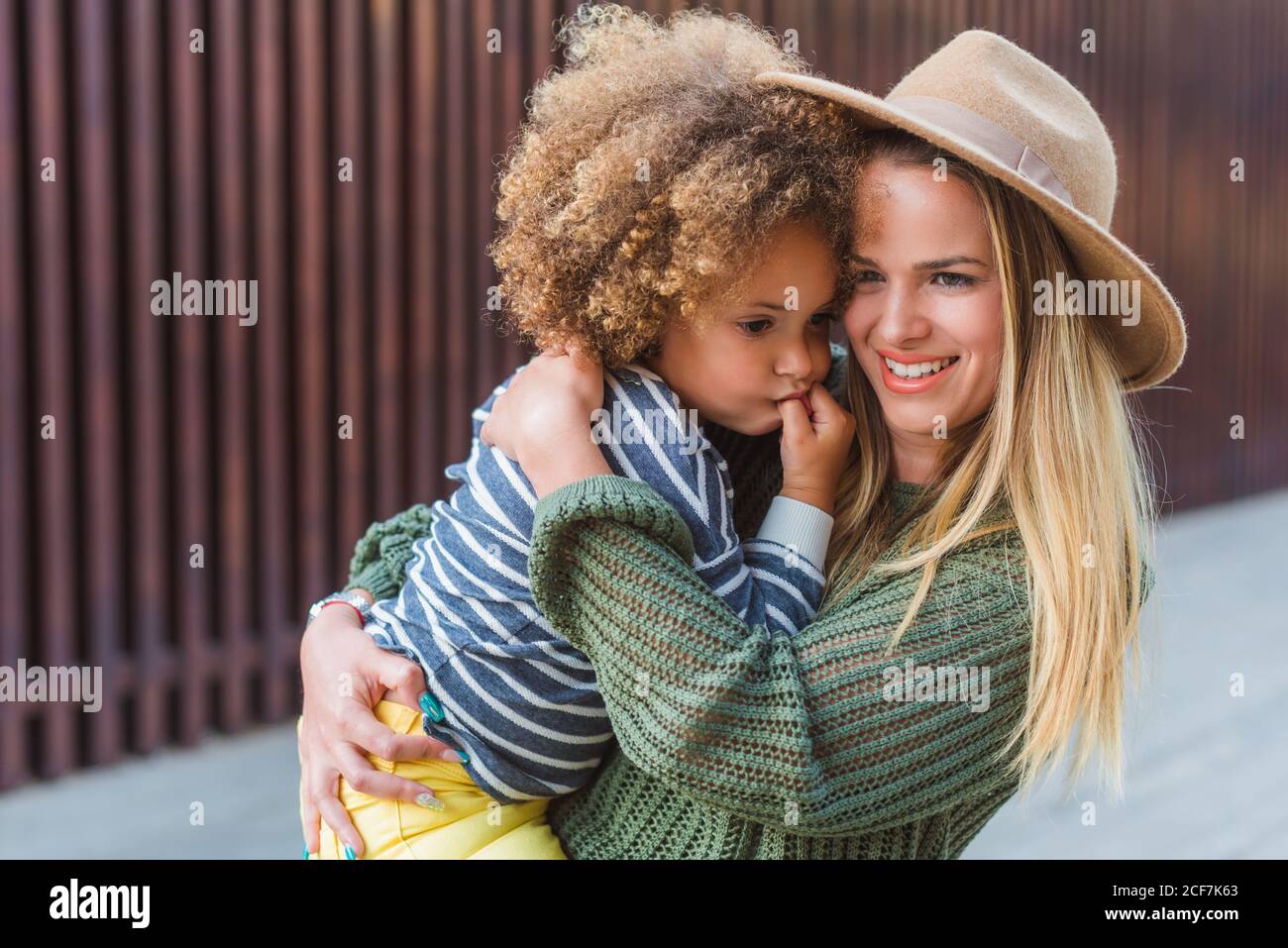 Vue latérale d'une jeune femme gaie dans un chandail tendance et chapeau enveloppant petite fille mignonne en se tenant dans la rue à proximité clôture moderne Banque D'Images