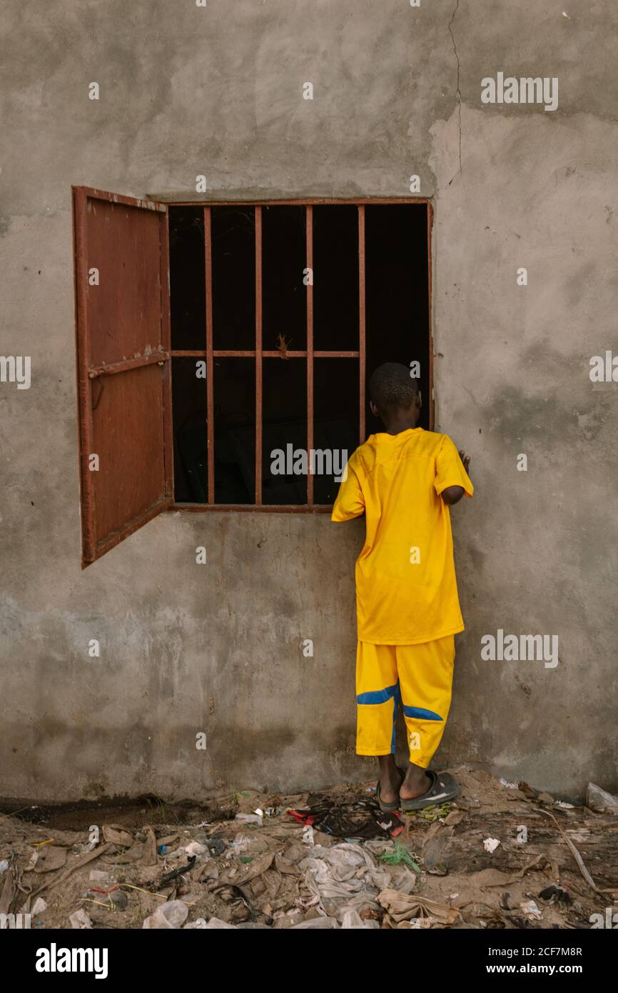 Gambie, Afrique - 3 août 2019 : vue arrière d'un garçon africain dans un costume jaune vif peeking à l'intérieur de la fenêtre sombre avec treillis de l'ancien bâtiment debout sur la terre des ordures Banque D'Images