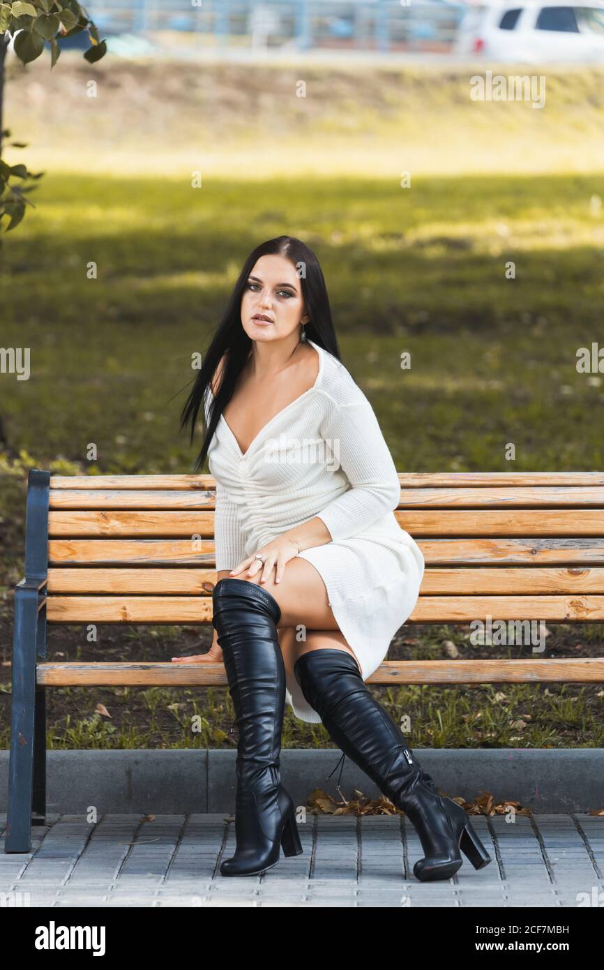 Jeune belle femme à cheveux foncés avec une silhouette élancée est effectivement assis sur un banc en robe blanche, de hauts talons contre le parc de la ville de freen Banque D'Images