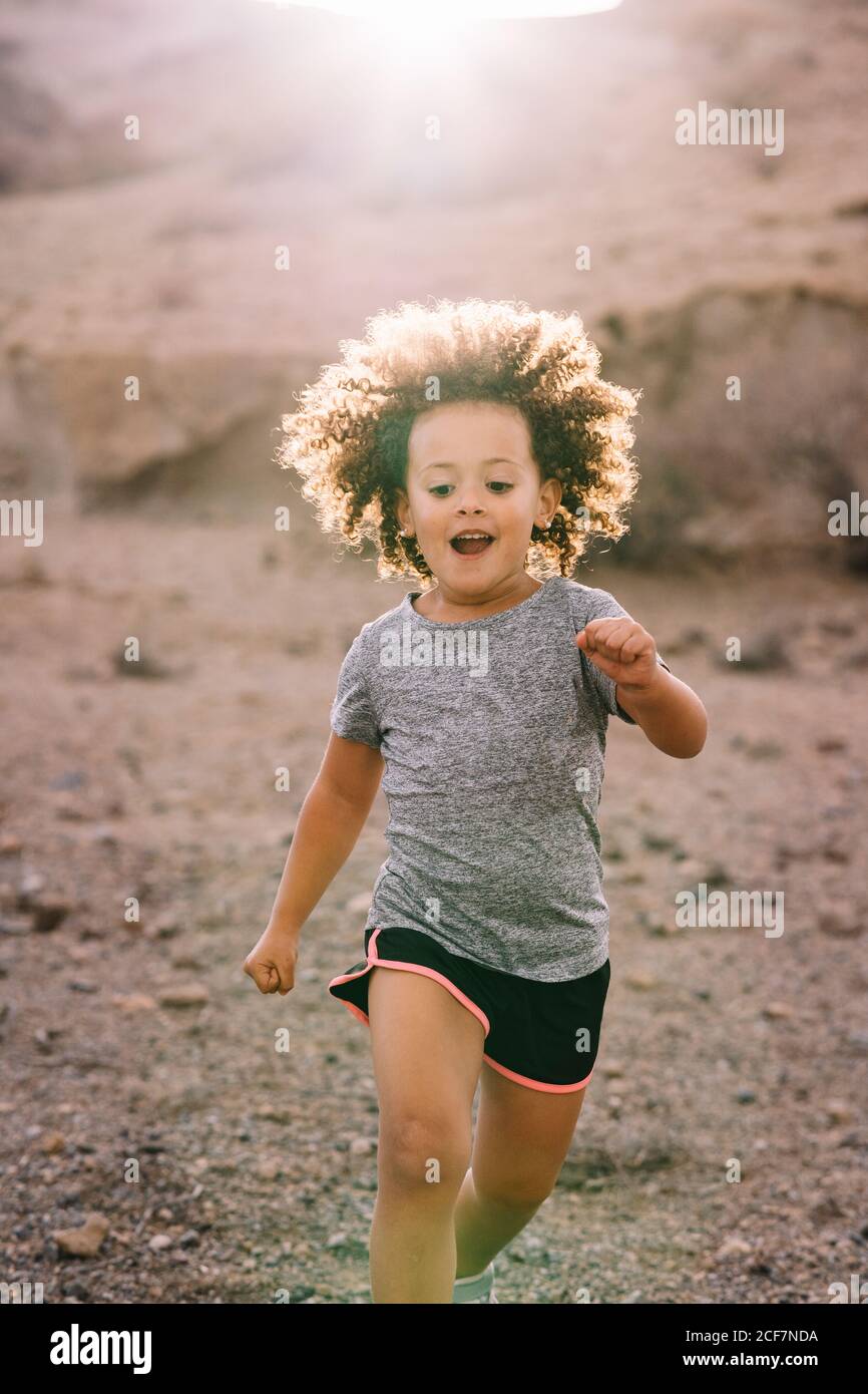 Joyeux enfant actif avec des cheveux bruns bouclés vêtus de sportive vêtements de jogging le matin sur un paysage sablonneux en contre-jour Banque D'Images