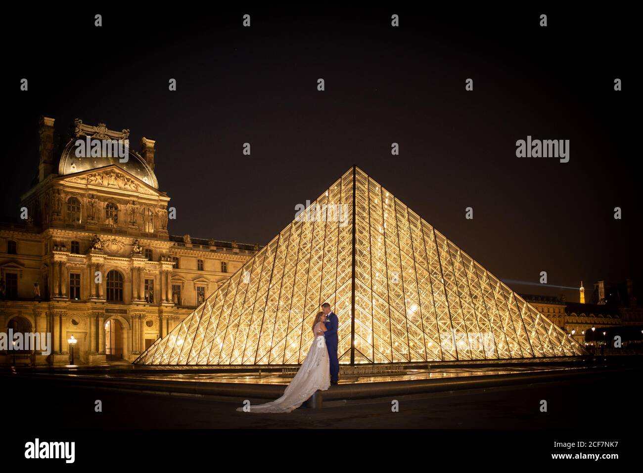 Jeune couple récemment marié en costume de mariage et robe embrassant Tout en se tenant dans une arche rockée avec le Louvre sur l'arrière-plan à Paris Banque D'Images