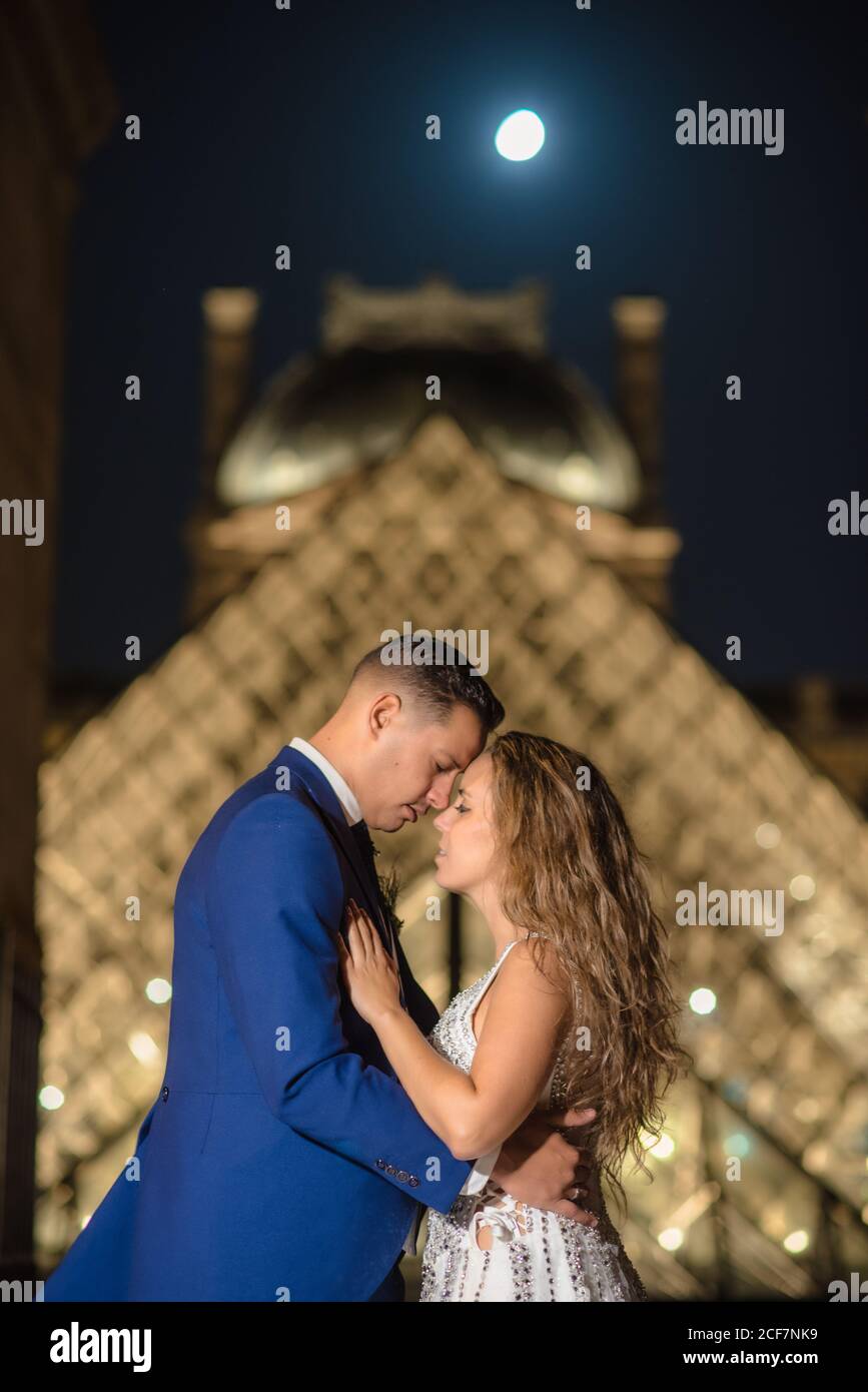Jeune couple récemment marié en costume de mariage et robe embrassant Tout en se tenant dans une arche rockée avec le Louvre sur l'arrière-plan à Paris Banque D'Images