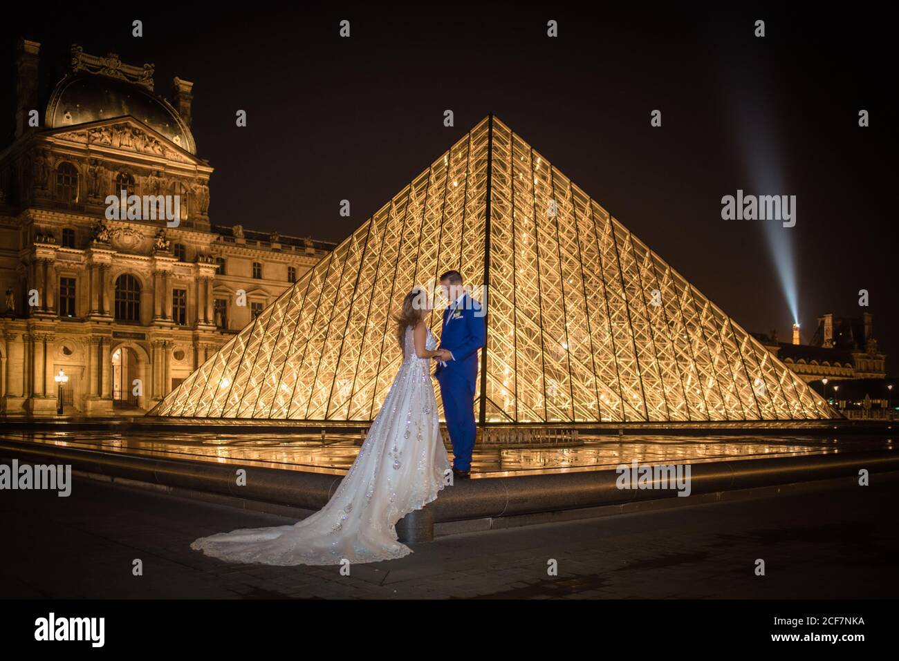 Jeune couple récemment marié en costume de mariage et robe embrassant Tout en se tenant dans une arche rockée avec le Louvre sur l'arrière-plan à Paris Banque D'Images