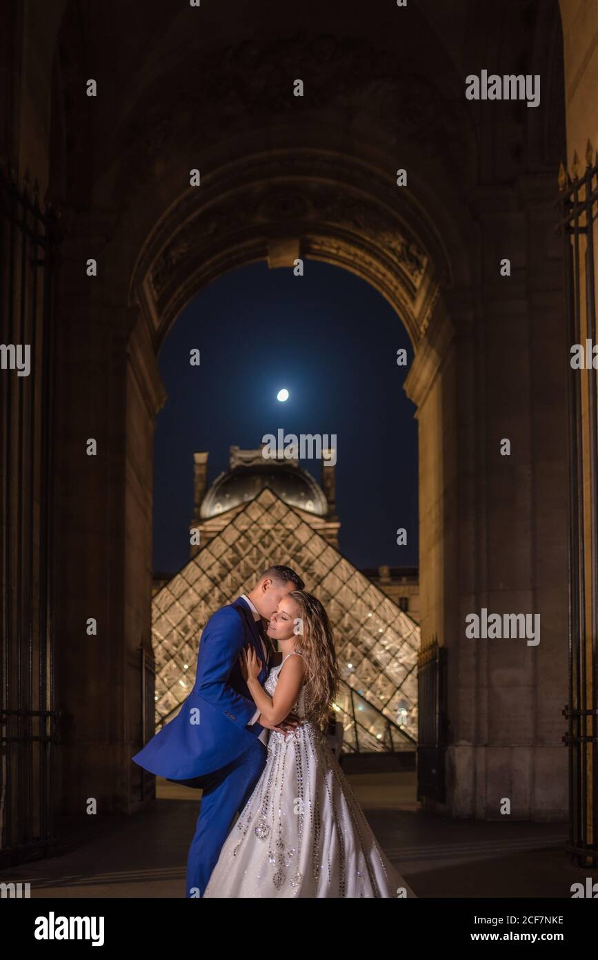 Jeune couple récemment marié en costume de mariage et robe embrassant Tout en se tenant dans une arche rockée avec le Louvre sur l'arrière-plan à Paris Banque D'Images